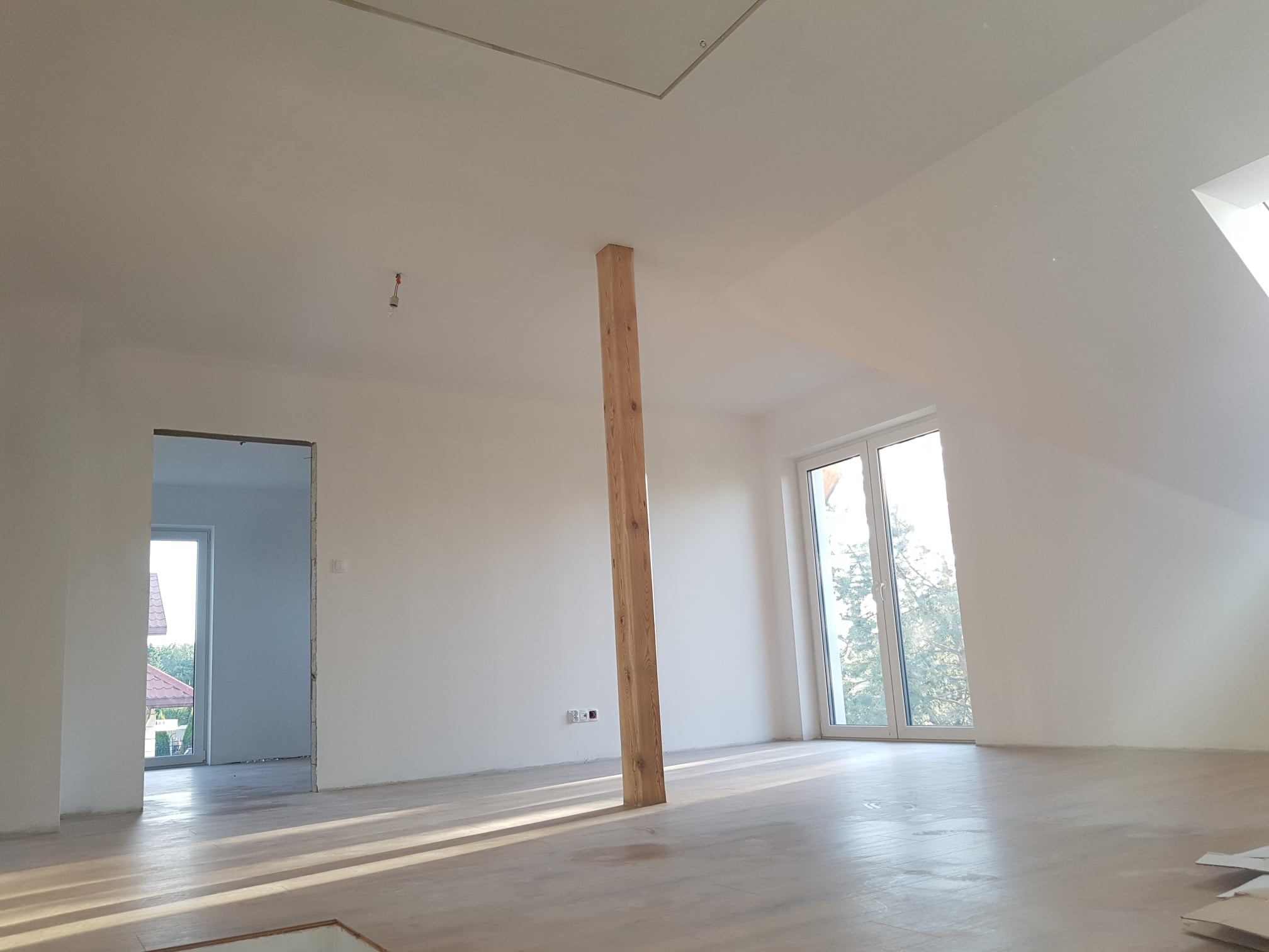 Poddasze w stanie deweloperskim z drewnianą kolumną wspierającą, białe ściany, jasna drewniana podłoga i okno dachowe wpadające w jasne światło.