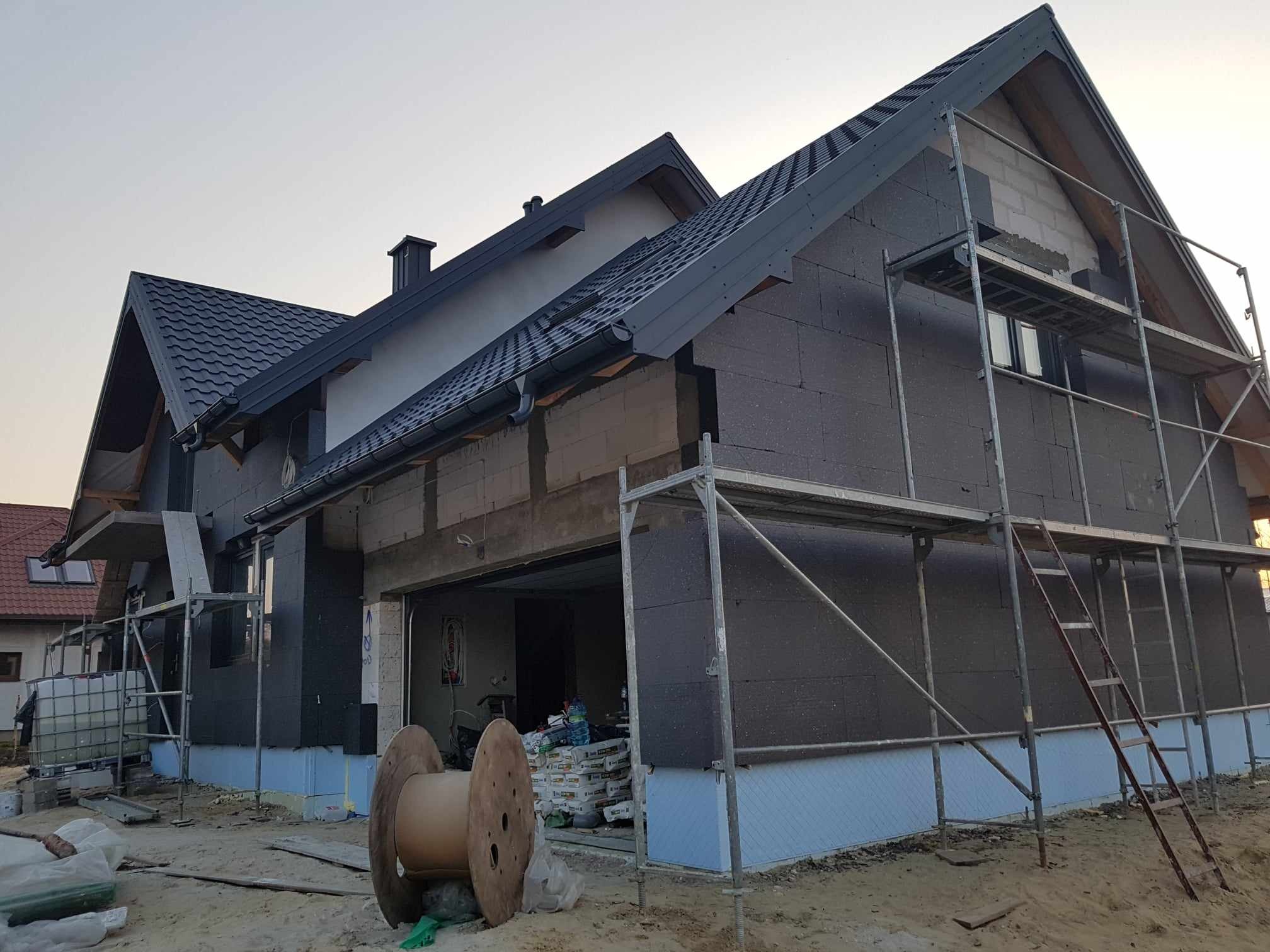 Dom w trakcie ocieplania zewnętrznego, widoczne rusztowania, rolki kablowe i worki z materiałami budowlanymi w otwartym garażu.