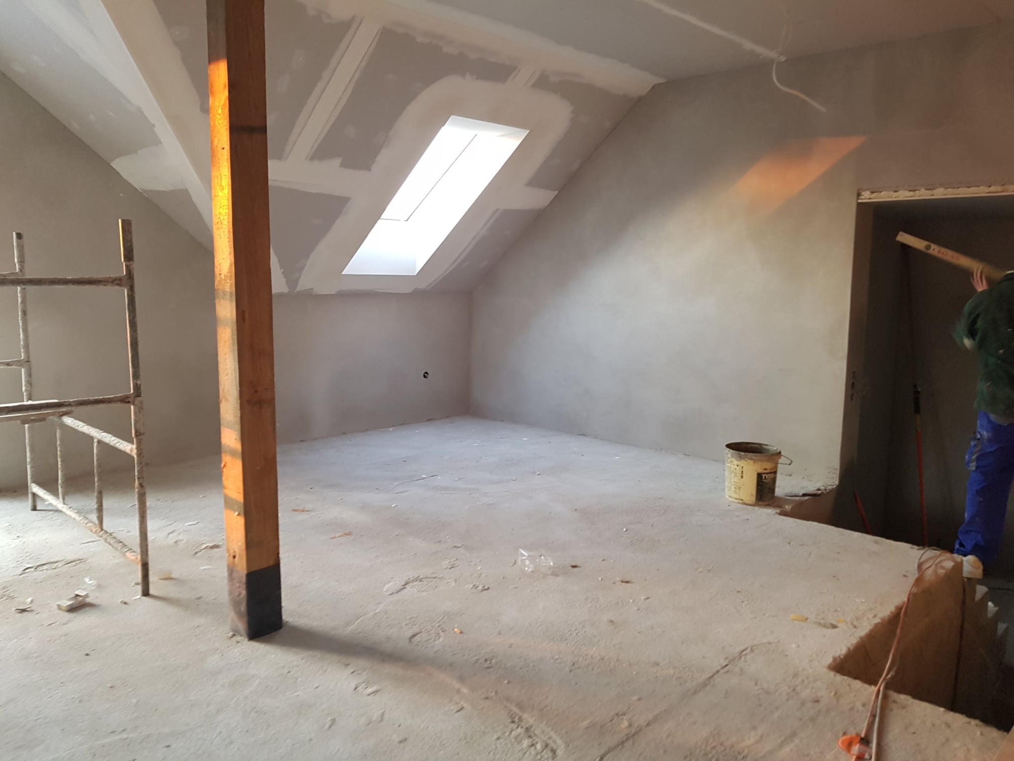Poddasze w trakcie wykańczania: widoczne szpachlowane ściany, okno dachowe, drewniany słup podtrzymujący, drabina malarska i pracownik z łatą w tle.