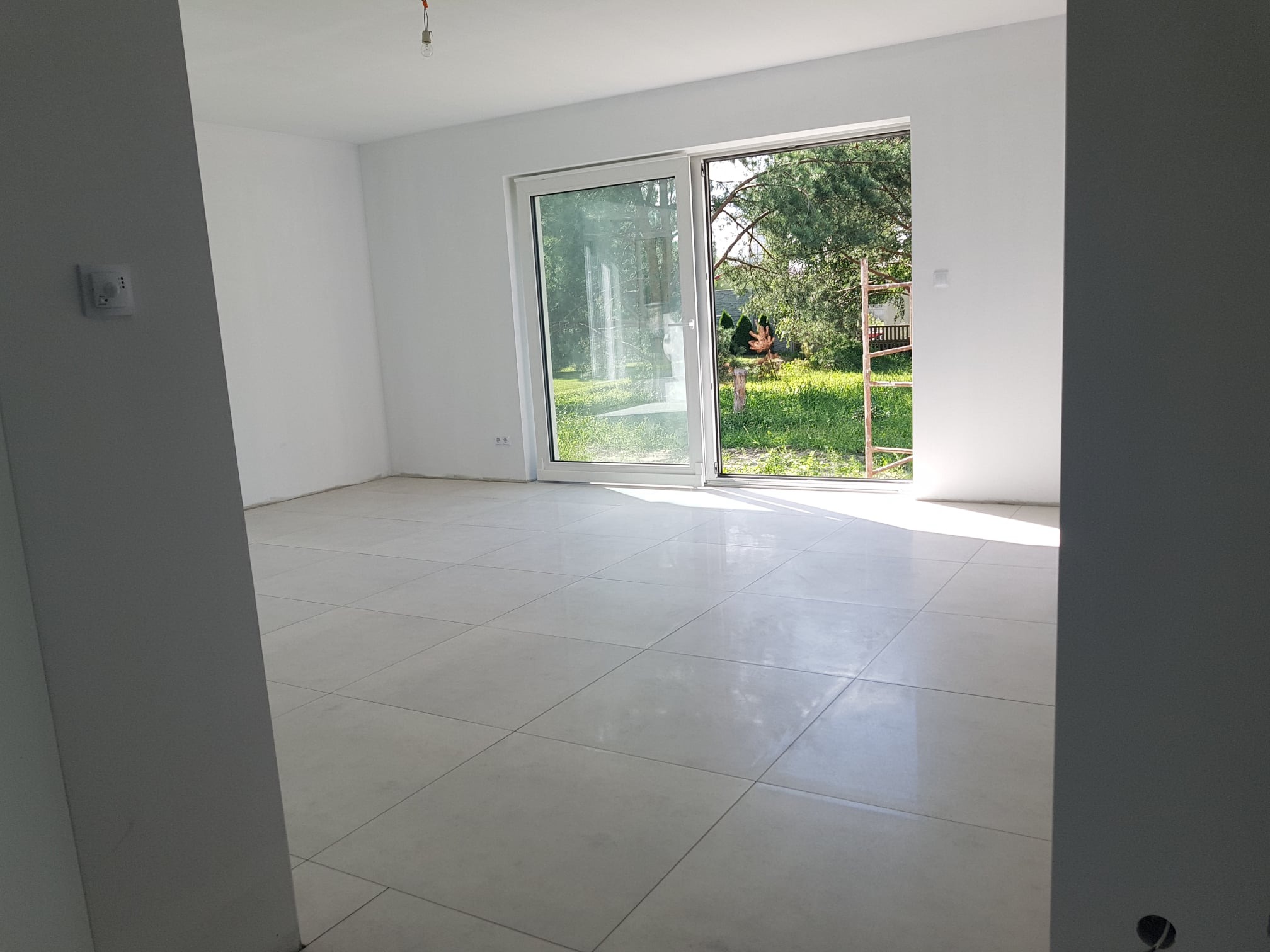 Wnętrze jasnego, nowo wykończonego pokoju z białymi ścianami, dużymi przesuwnymi drzwiami balkonowymi wychodzącymi na ogród, i podłogą wyłożoną jasnymi, kwadratowymi płytkami ceramicznymi.