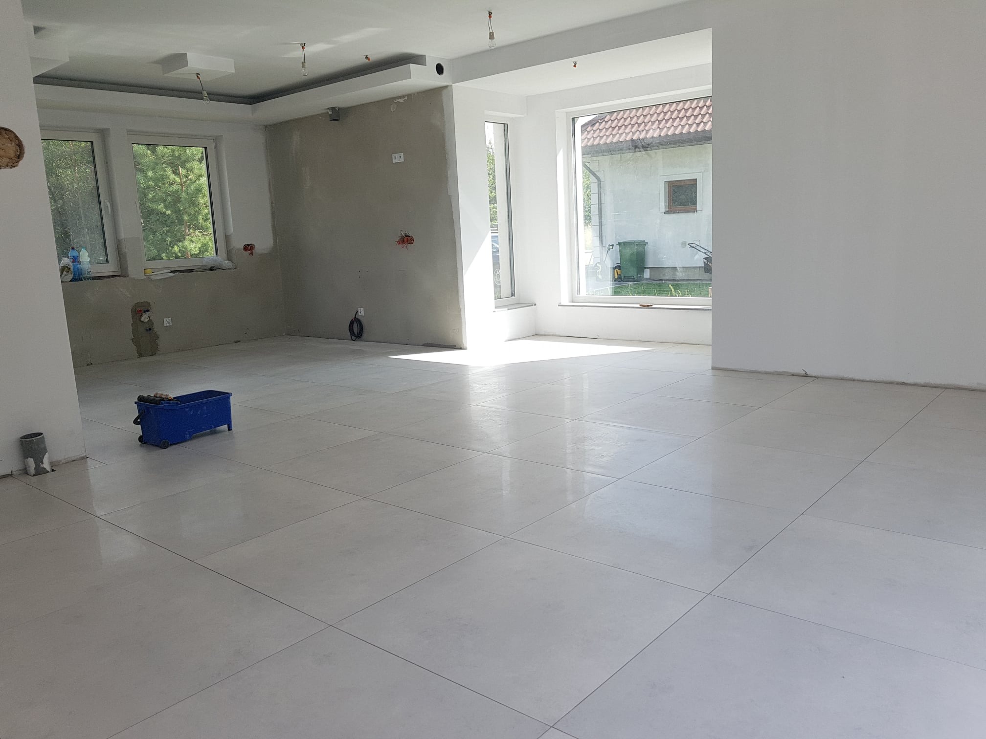 Jasne, duże płytki podłogowe w nowym, przestronnym pomieszczeniu z dwoma oknami, ściany częściowo w stanie surowym, niebieski wózek z narzędziami w rogu, widoczne elementy instalacji elektrycznej.