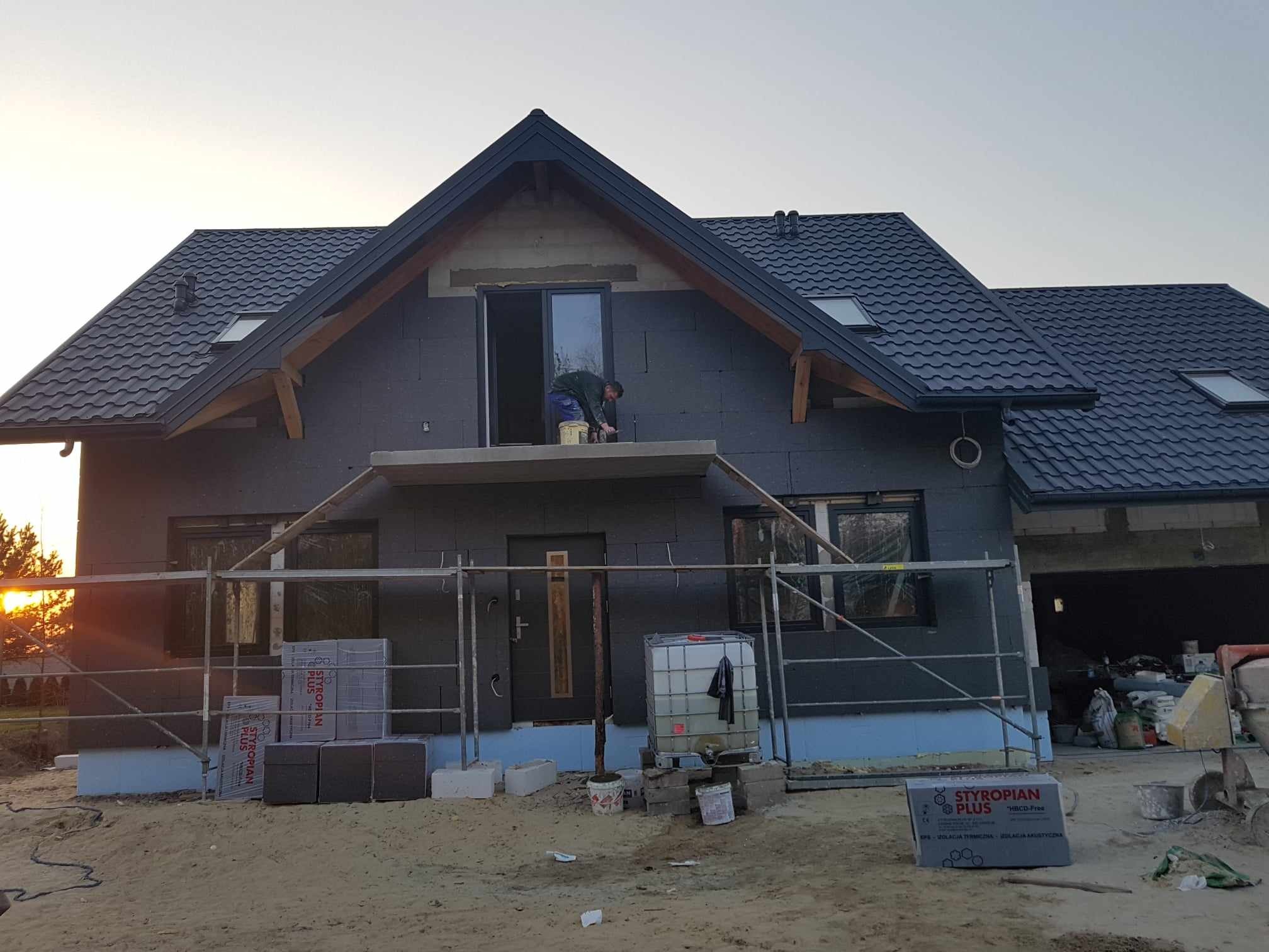 Dom murowany w trakcie ocieplania styropianem grafitowym, widoczne rusztowania i pracownik na balkonie, przygotowujący się do nałożenia warstwy wykończeniowej.