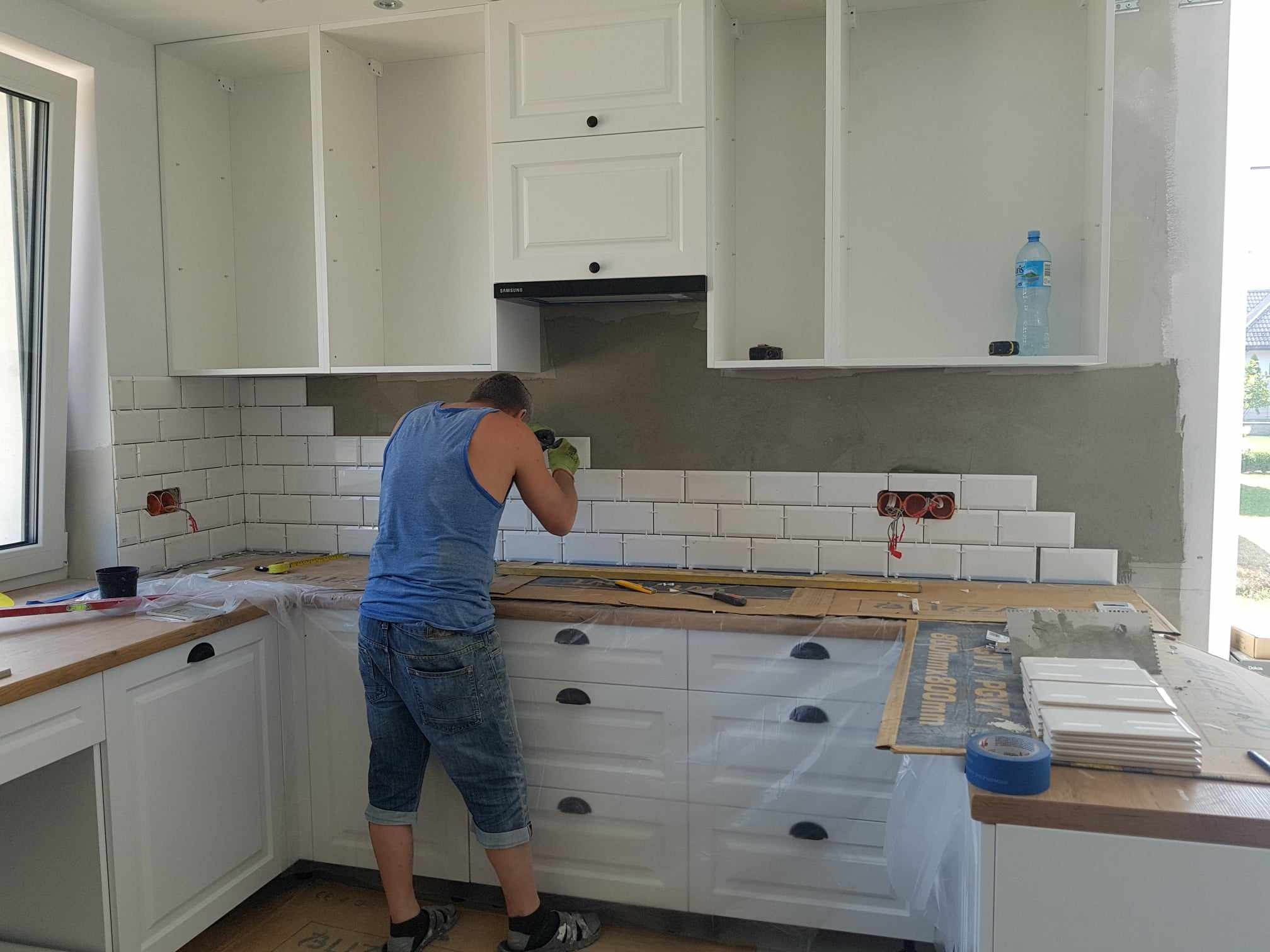 Układanie białych, prostokątnych płytek na ścianie w kuchni; widoczny glazurnik w niebieskim podkoszulku, częściowo wykończone szafki kuchenne w stylu rustykalnym i narzędzia pracy.