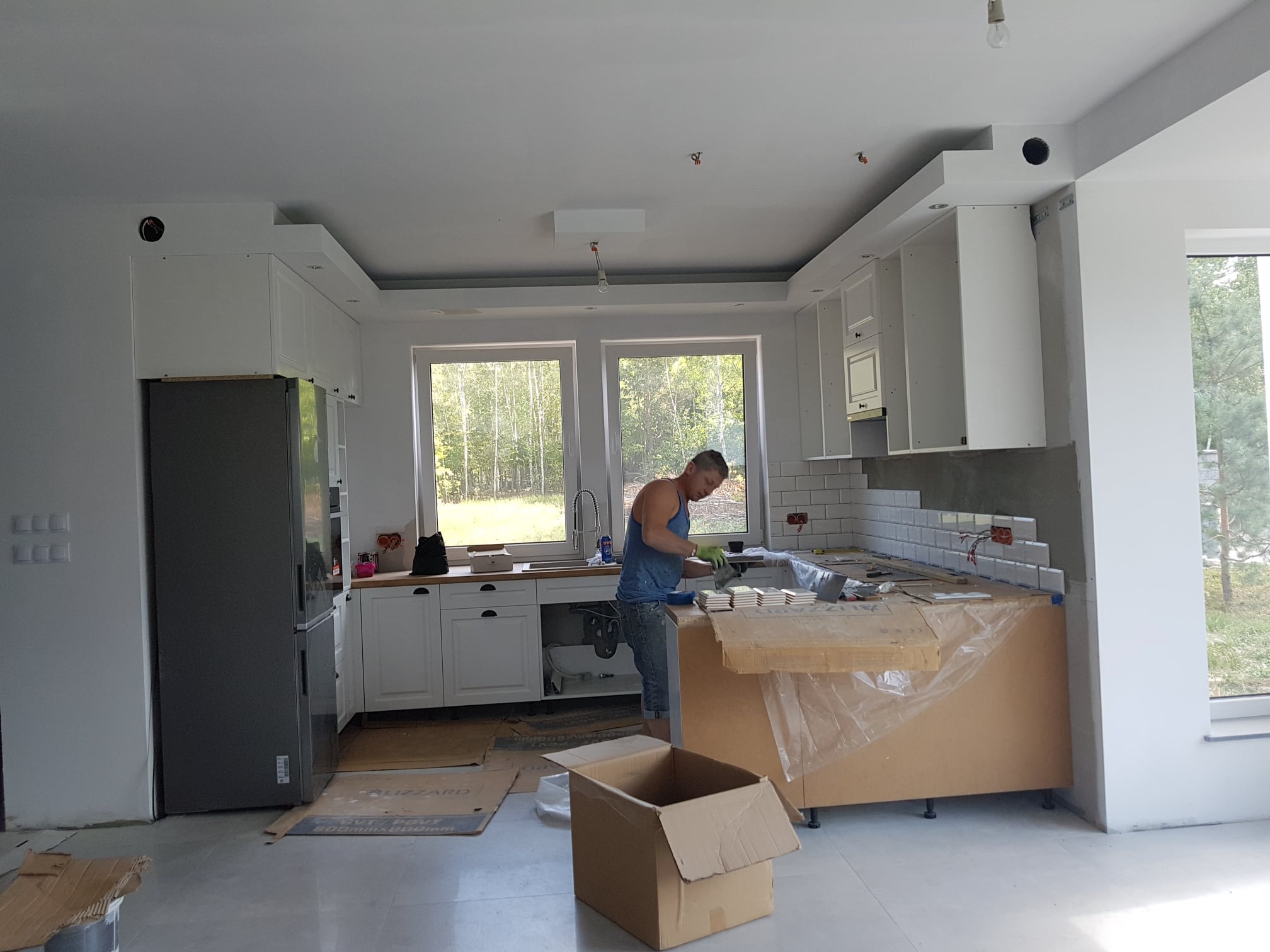 Mężczyzna w kuchni, w trakcie układania białych, prostokątnych płytek na ścianie nad blatem. Częściowo wykończona ściana, widoczne narzędzia, kartony z płytkami i niedokończone szafki kuchenne.