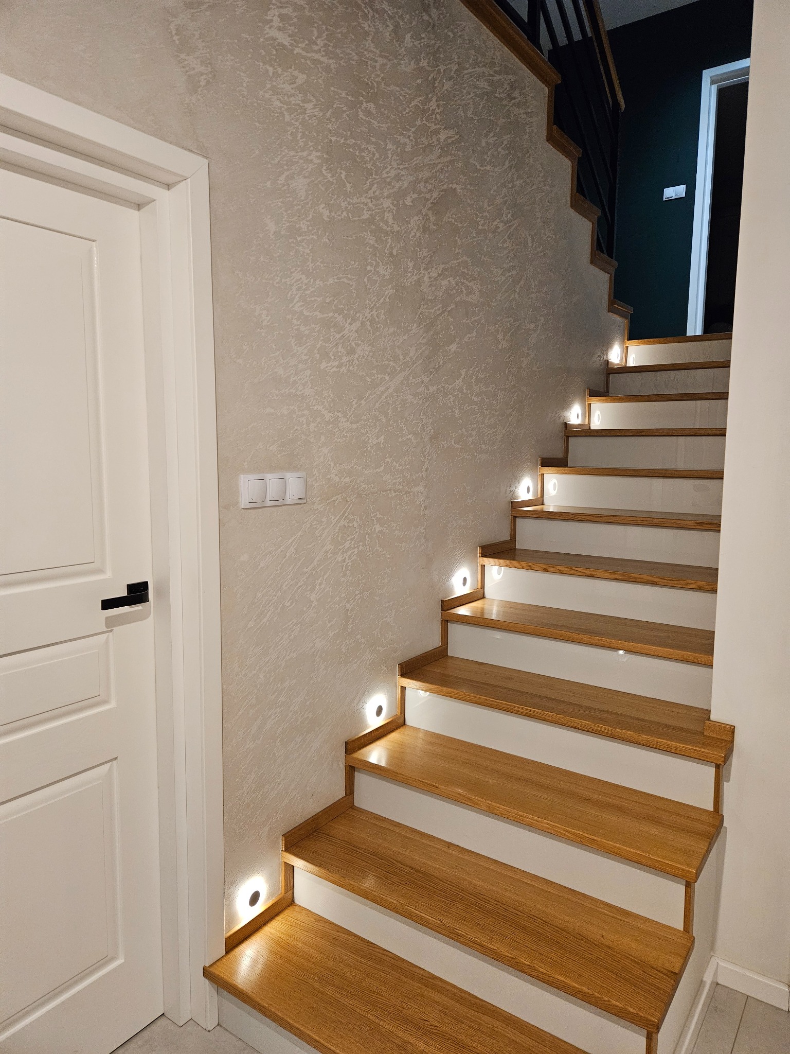 Nowoczesne schody drewniane z białymi podstopnicami i subtelnym oświetleniem LED w ścianie o dekoracyjnej fakturze, obok białych drzwi z czarną klamką.
