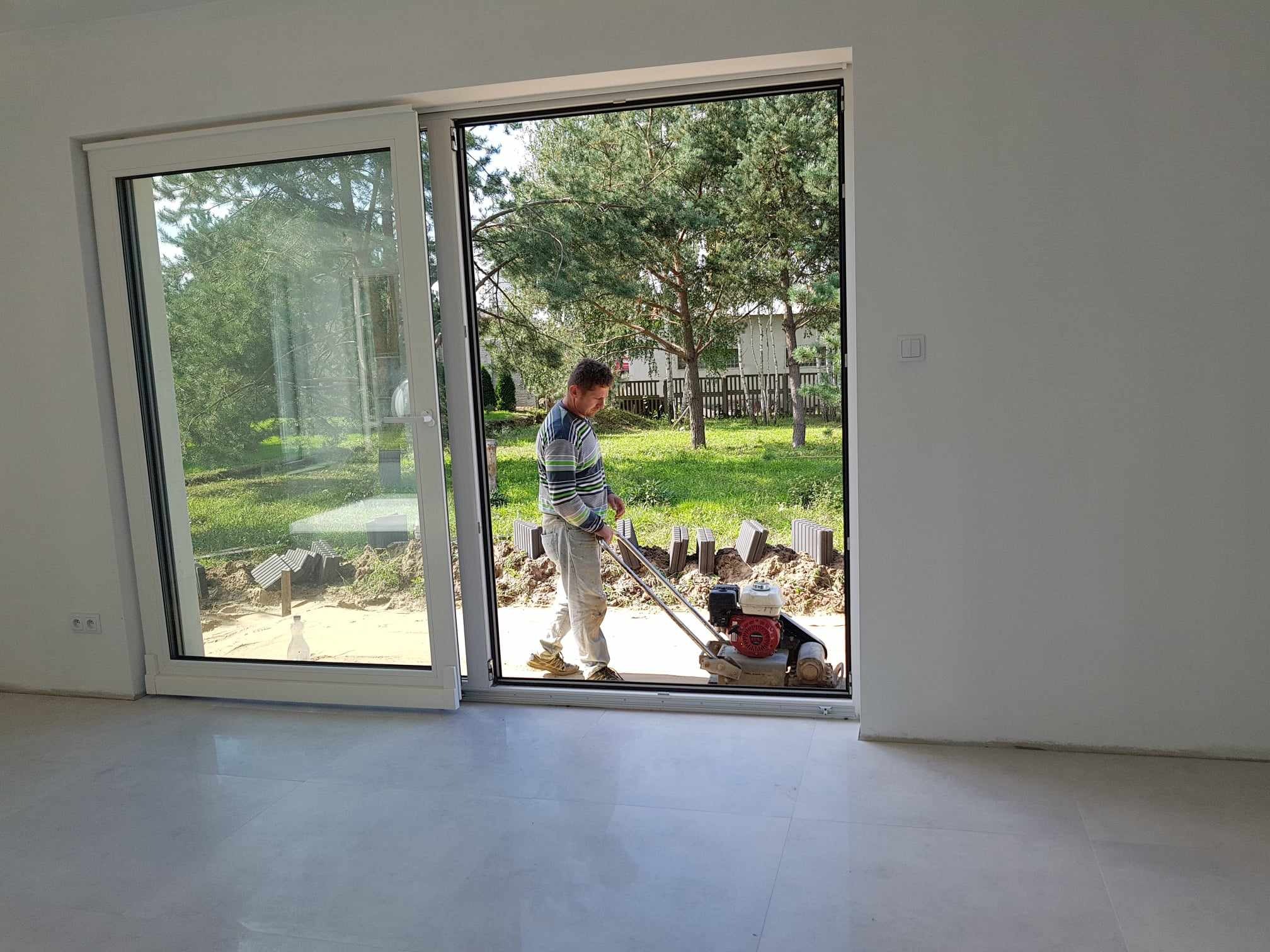 Pracownik zagęszcza grunt podłoża pod taras przed dużym oknem przesuwnym nowo wykończonego domu, widoczne ułożone krawężniki i butelka wody.