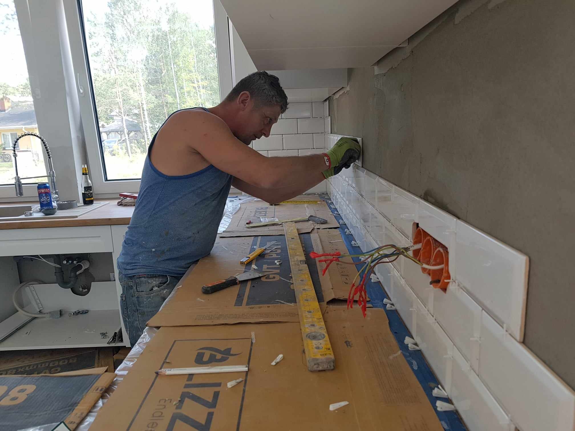 Mężczyzna w trakcie układania białych, prostokątnych płytek nad blatem kuchennym, widoczne puszki elektryczne i poziomica na zabezpieczonym kartonem blacie.