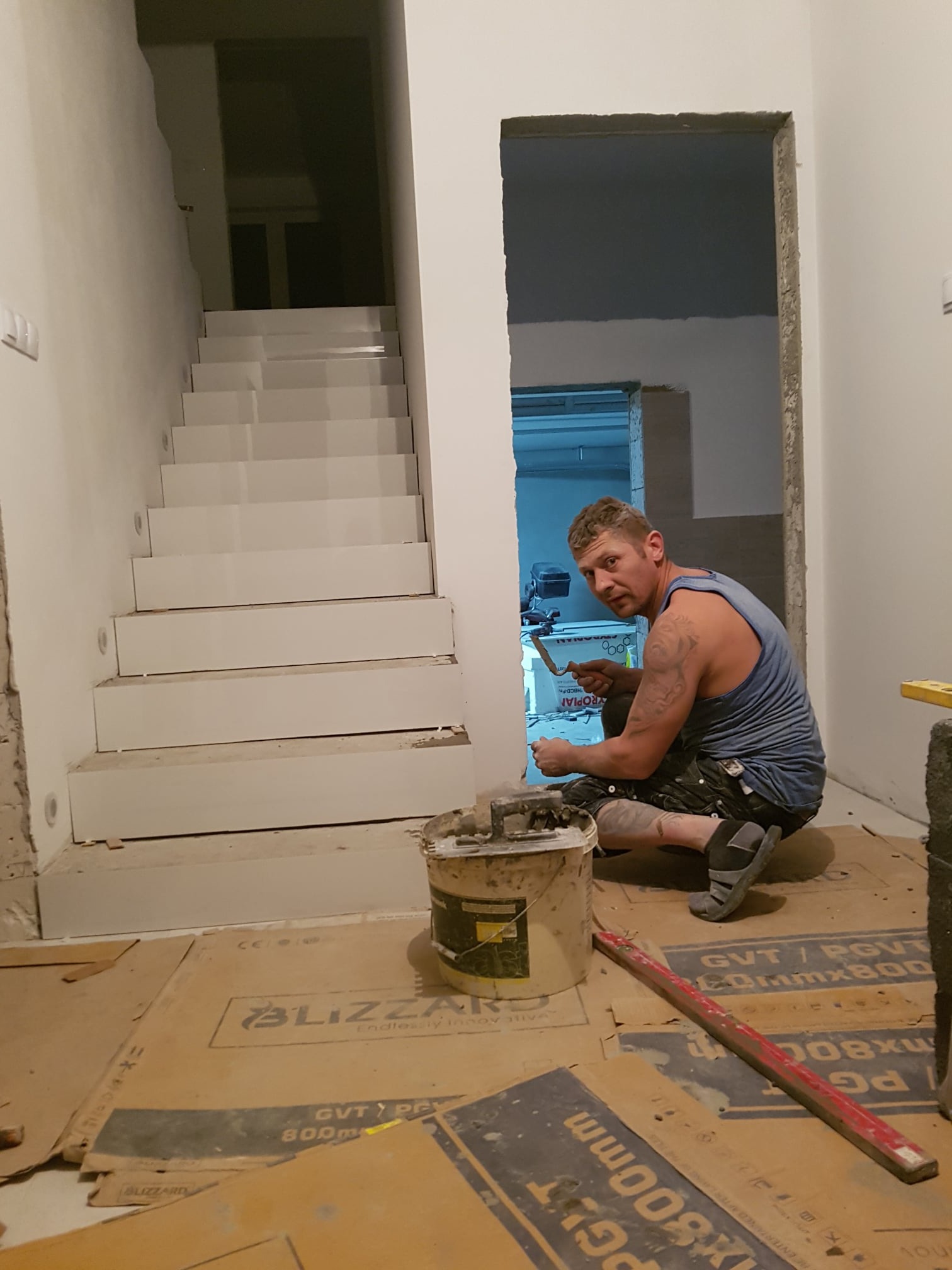 Mężczyzna w niebieskiej koszulce na ramiączkach, klęczy na podłodze zabezpieczonej kartonem, wygładzając zaprawę szpachelką wokół ościeżnicy, widoczne białe schody w tle.