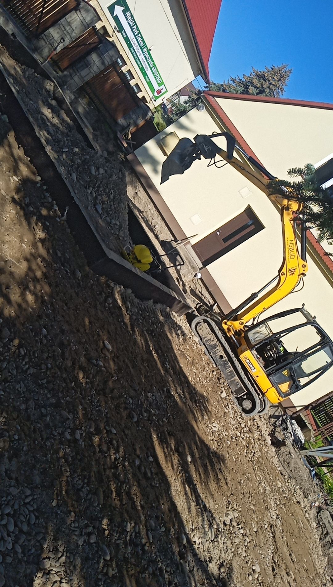 Żółta mini koparka JCB na gąsienicach, obok wykopu z widocznym żółtym zagęszczarką gruntu, na tle budynku z brązowymi drzwiami i tablicą informacyjną o Miejskim Parku Zieleni.