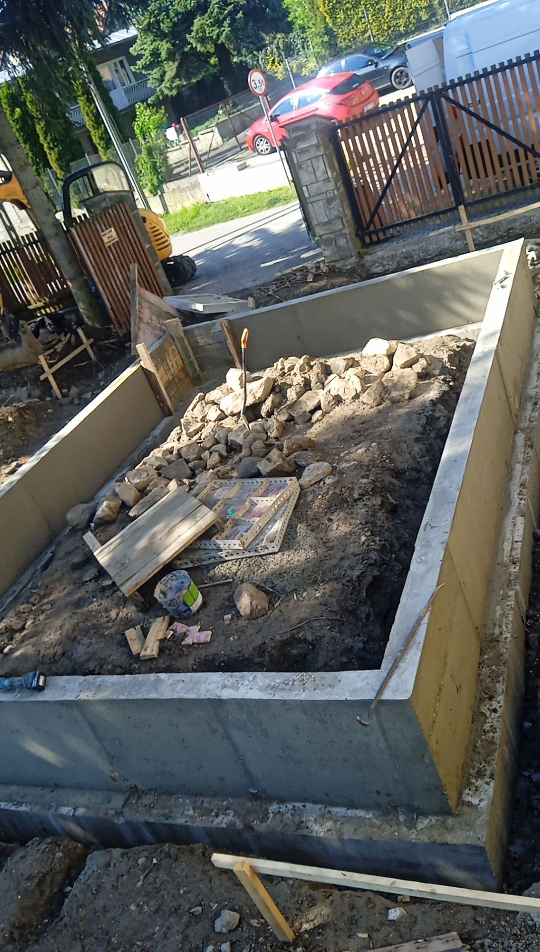 Przygotowany wykop z betonowymi ścianami i częściowo wypełniony gruzem kamiennym, widoczne narzędzia i sprzęt budowlany w tle, sugerujący etap prac ziemnych.