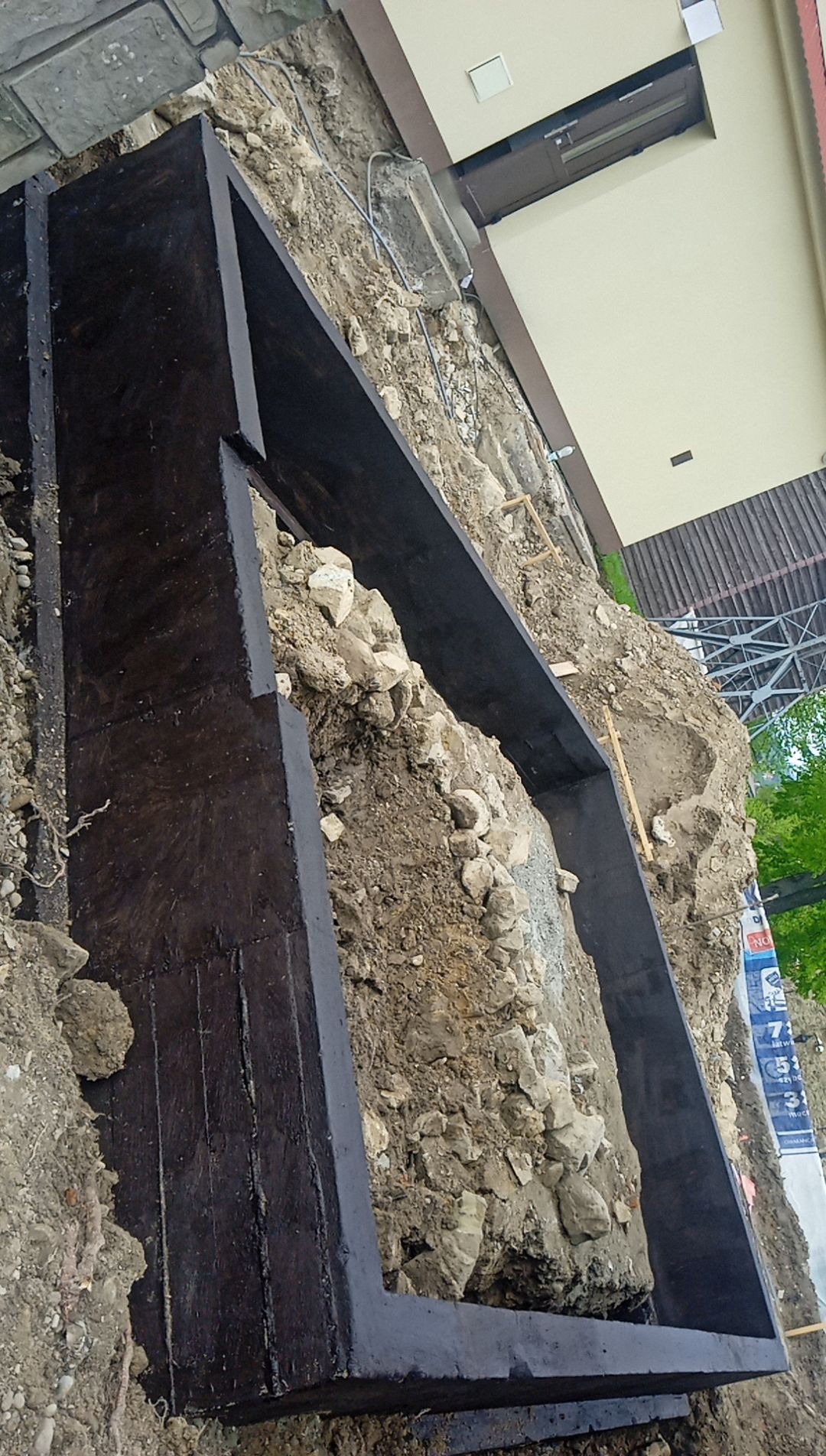 Wykop pod fundament z deskowaniem w trakcie prac ziemnych, widoczne kamienie i glina, budynek w tle.
