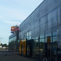 Jarosław Dembecki Malar-Tech - Elewacja budynku salonu samochodowego Opel w trakcie czyszczenia lub malowania; widoczne rusztowania, folie ochronne i elementy reklamowe.