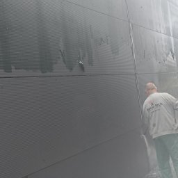 Jarosław Dembecki Malar-Tech - Pracownik w uniformie firmy Malar-tech myje ciśnieniowo szarą, panelową elewację przemysłową, widoczne ślady zabrudzeń na powierzchni.