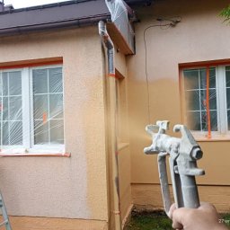Jarosław Dembecki Malar-Tech - Natryskowe malowanie elewacji domu jednorodzinnego. Okna zabezpieczone folią, widoczny pistolet natryskowy i drabina. Częściowo pomalowana ściana w odcieniach beżu.
