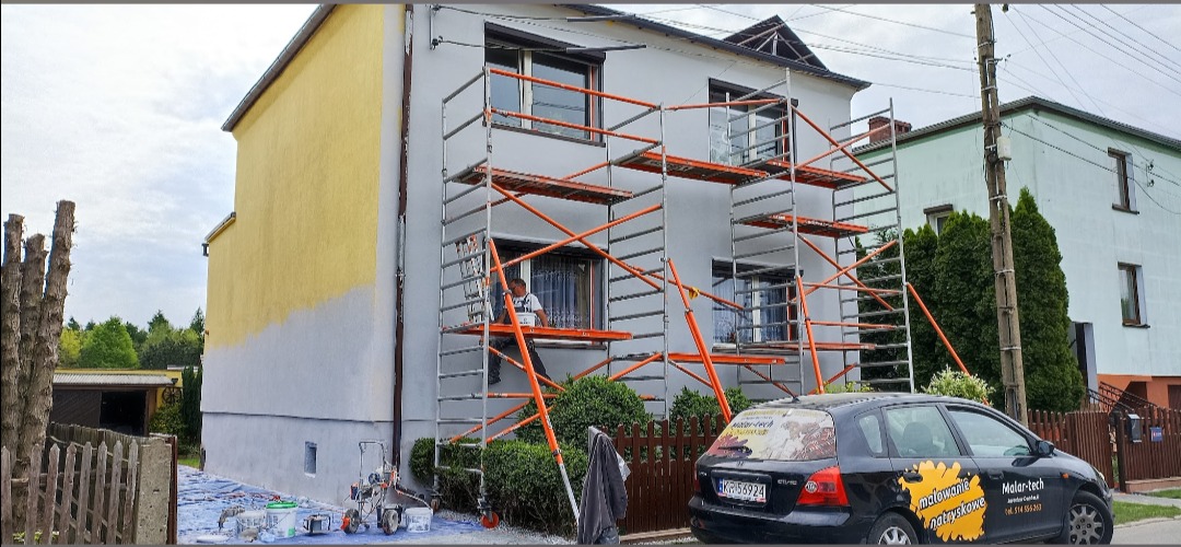 Dwupiętrowy dom w trakcie malowania elewacji na żółto, z rusztowaniem, pracownikiem w kombinezonie na rusztowaniu i samochodem firmy z reklamą malowania natryskowego w tle.