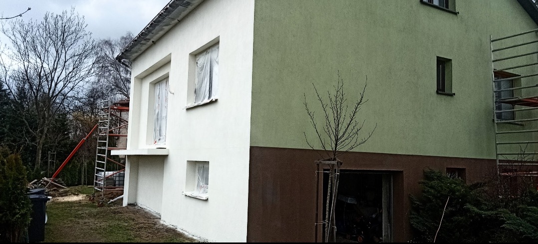 Dwukondygnacyjny dom z częściowo odnowioną elewacją; jedna ściana pomalowana na biało z oknami zabezpieczonymi folią, druga w odcieniach zieleni i brązu, z widocznym rusztowaniem i młodym drzewkiem.