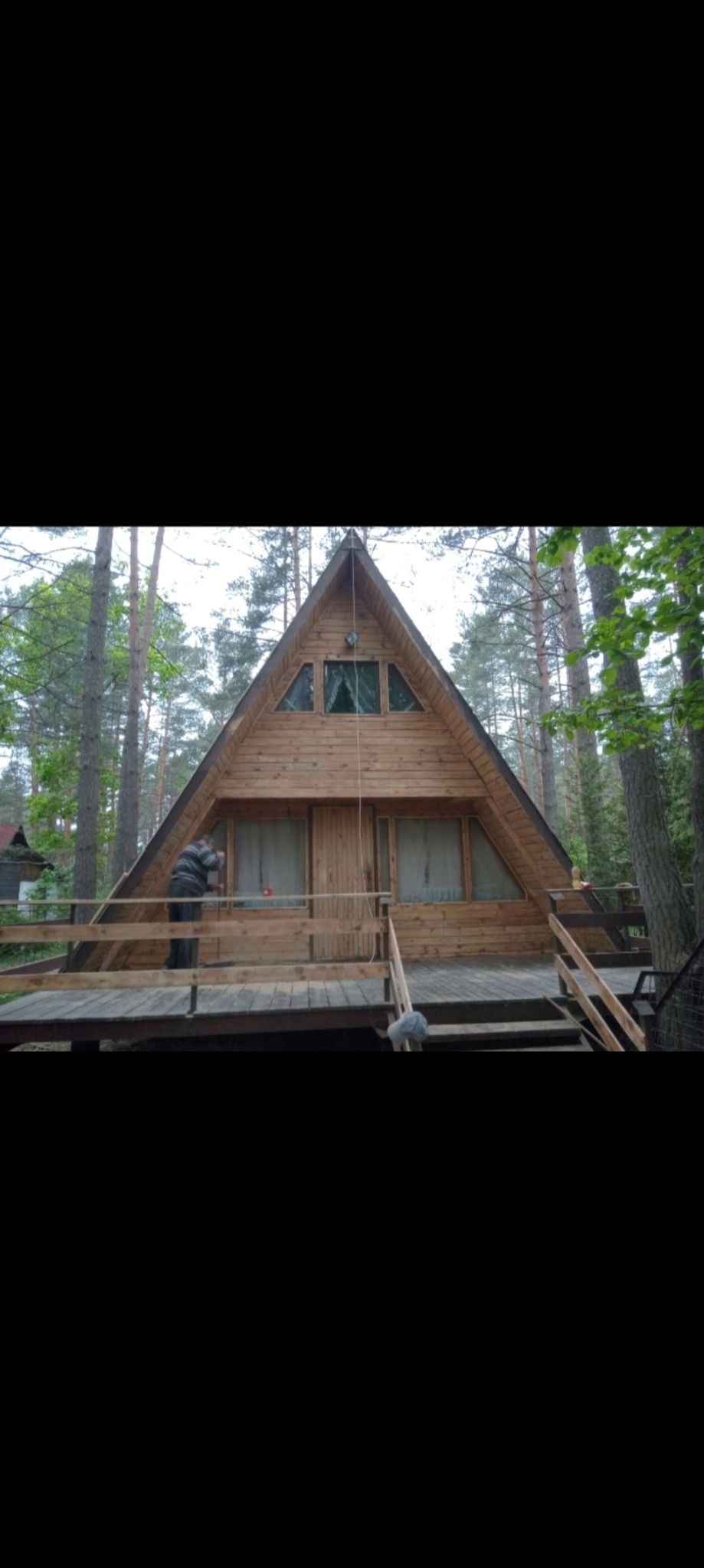 Drewniany dom w kształcie trójkąta na tle lasu, pracownik dokonuje konserwacji elewacji z drewnianych desek na tarasie.