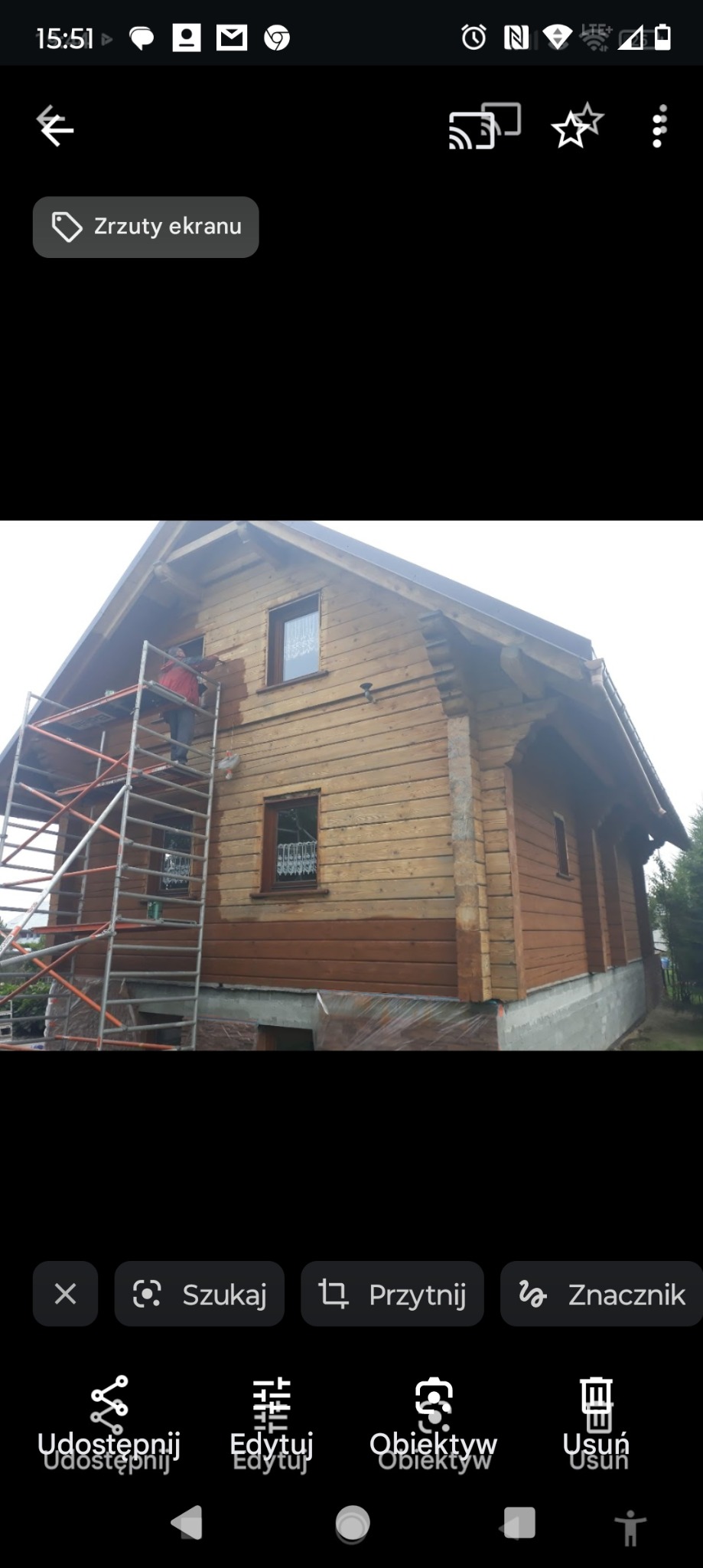 Drewniany dom w trakcie częściowego malowania elewacji na brązowo, widoczne rusztowanie i pracownik w czerwonej kurtce.