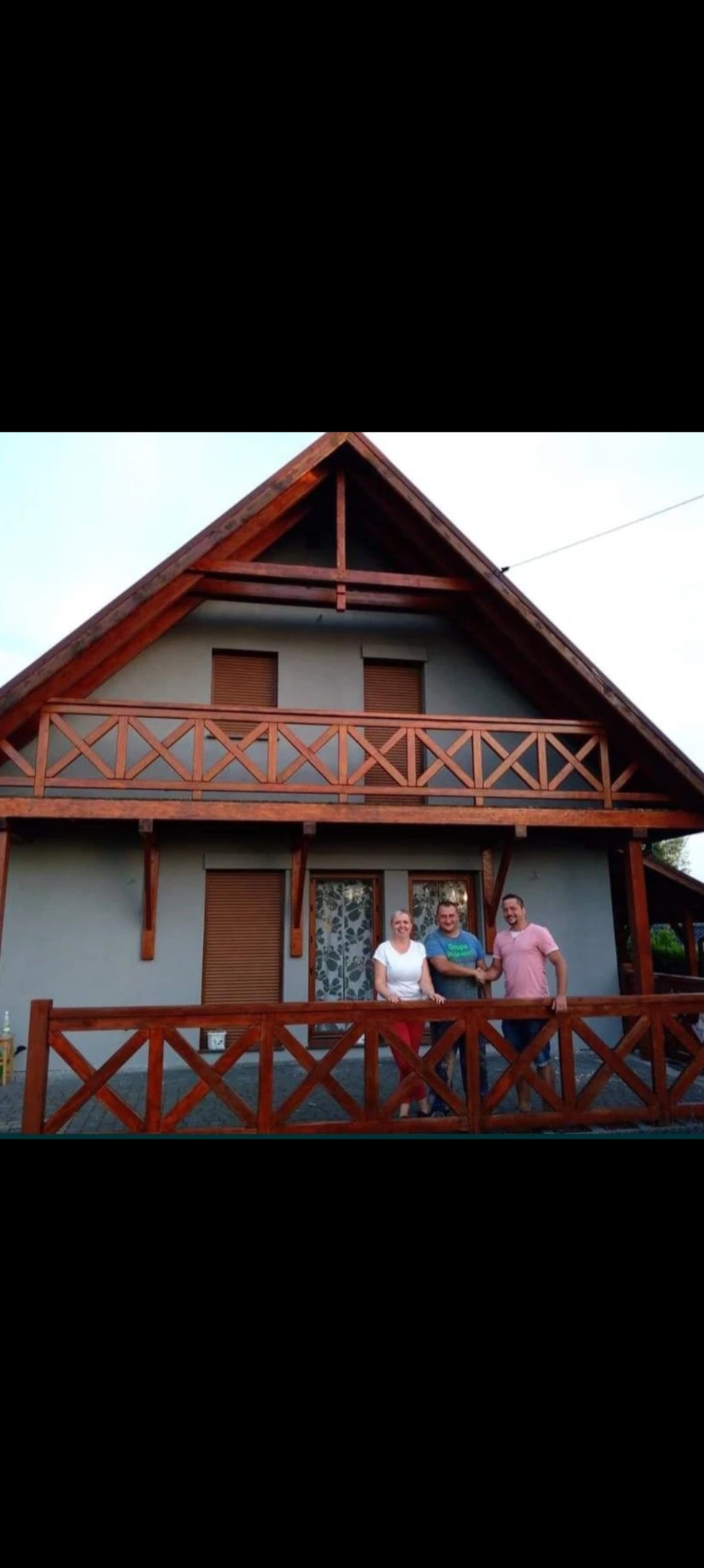 Szary dom jednorodzinny z drewnianą konstrukcją dachu i balustradami, na tle którego pozują trzy osoby.