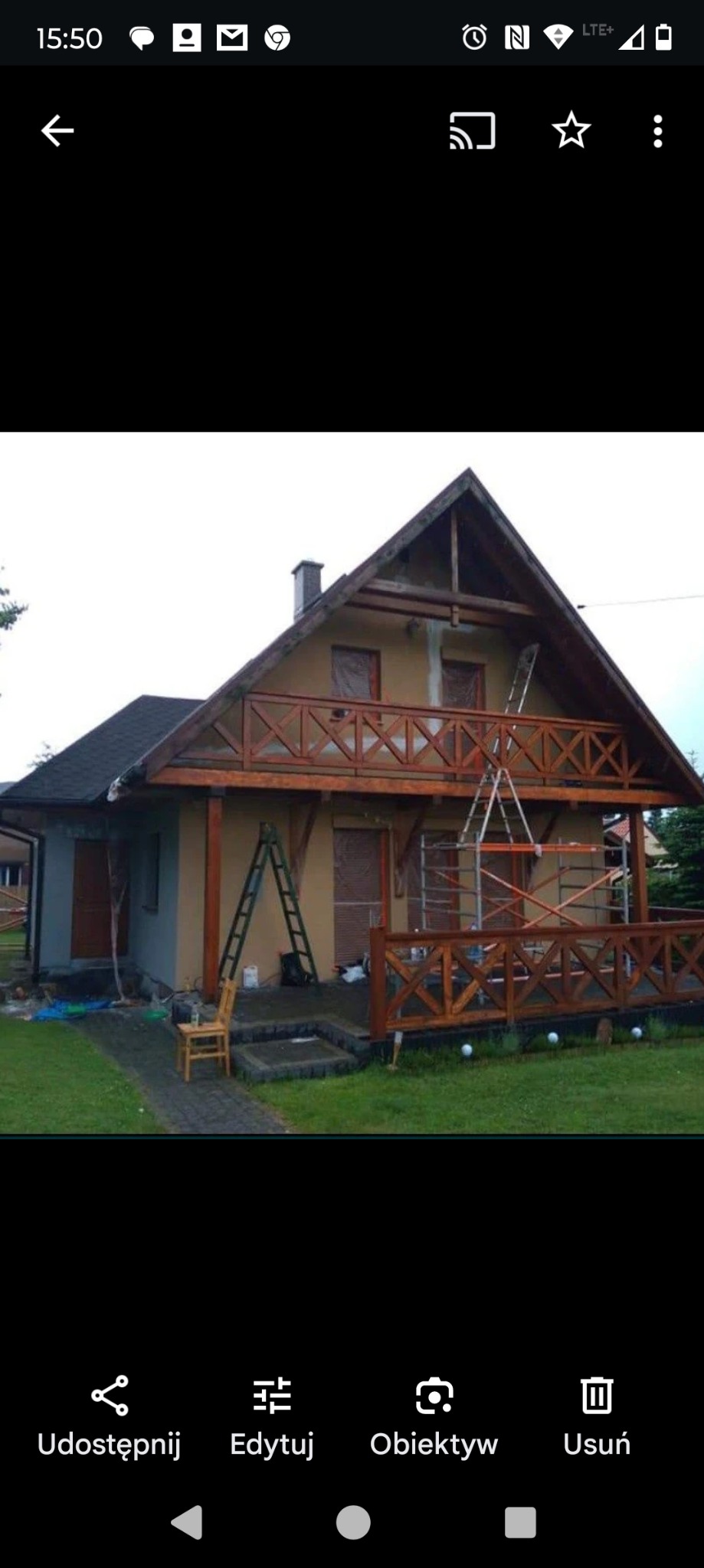 Dom z drewnianą konstrukcją balkonu, na elewacji widoczne ślady prac malarskich, drabina oparta o ścianę, rusztowanie wiszące na linie.