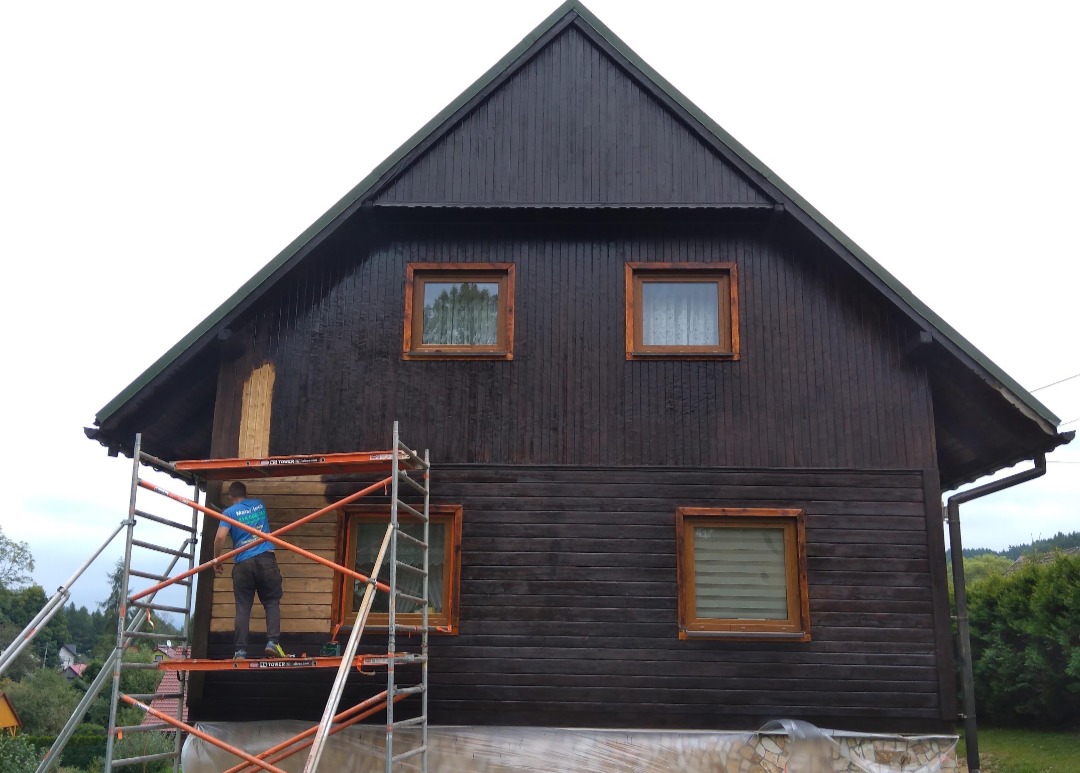 Dwukondygnacyjny dom z ciemnobrązową drewnianą elewacją w trakcie malowania; fragment elewacji jeszcze niepomalowany, pracownik na rusztowaniu w niebieskiej koszulce maluje ścianę.