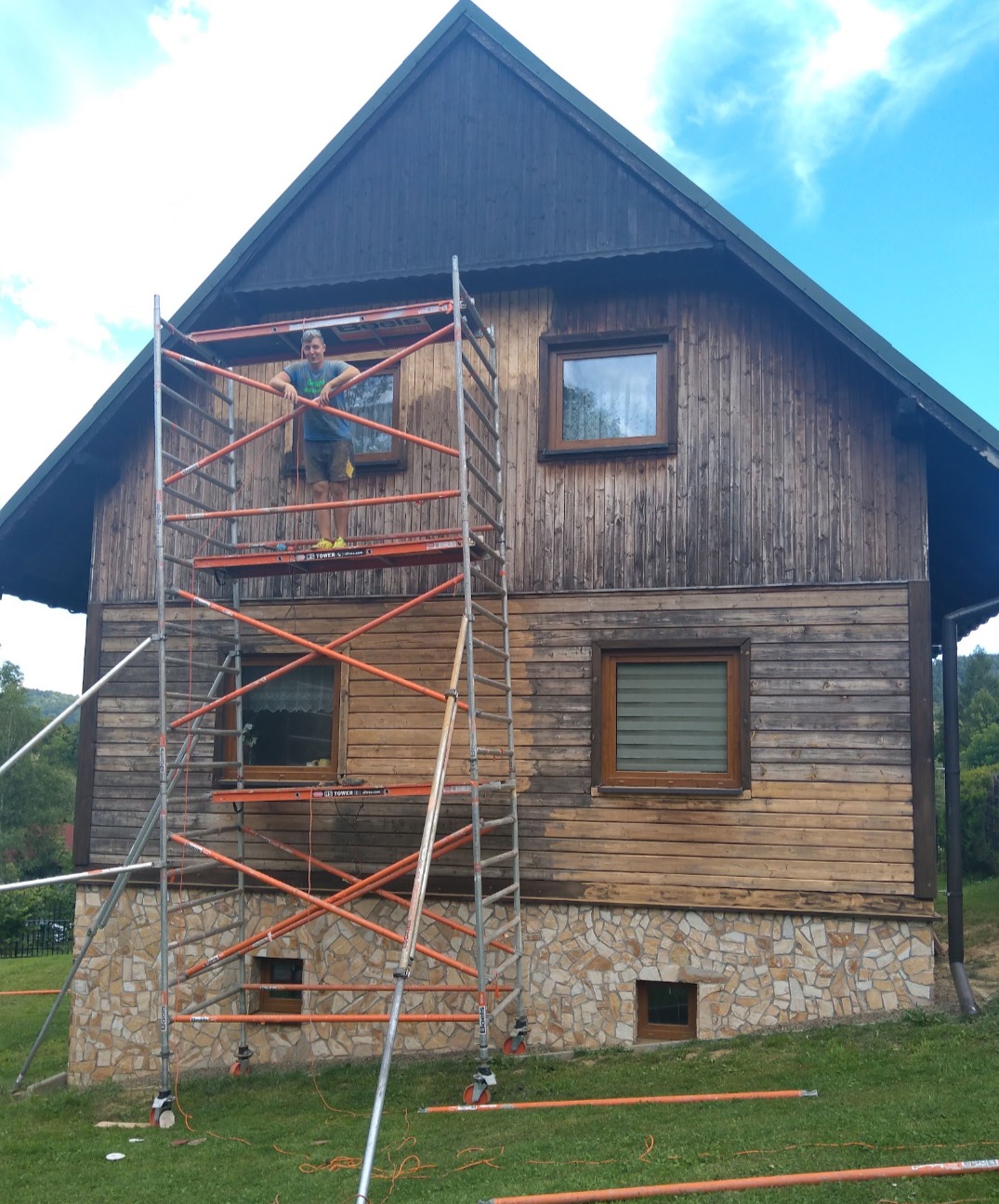 Drewniany dom z częściowo odnowioną elewacją, widoczny pracownik na rusztowaniu, fragmenty ściany w trakcie czyszczenia lub przygotowania do malowania, podmurówka z kamienia.