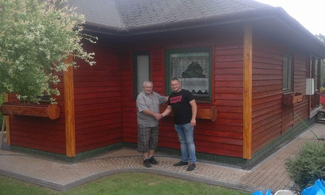 Dwaj mężczyźni ściskają sobie dłonie przed domem z drewnianą elewacją pomalowaną na czerwono-brązowy kolor, z widocznymi donicami pod oknami, kostka brukowa na ścieżce.