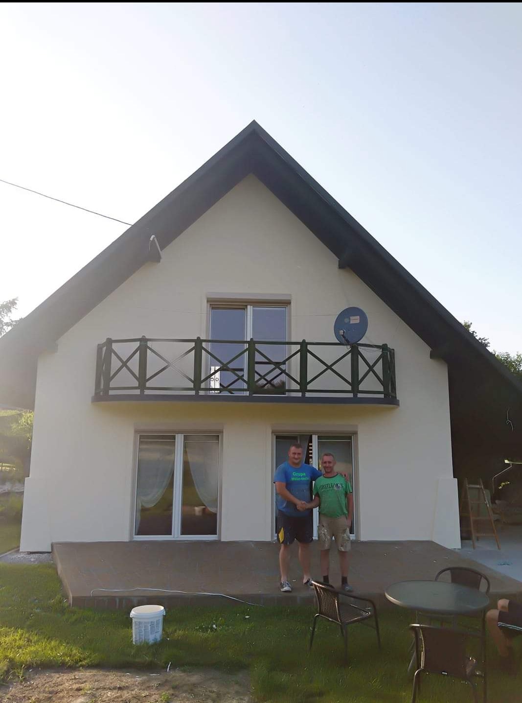 Dwóch mężczyzn ściska sobie dłonie na tle odnowionej elewacji domu z balkonem i anteną satelitarną, widoczny częściowo stół ogrodowy i wiadro z farbą.