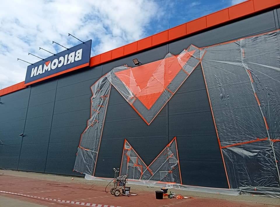 Malowanie elewacji budynku z logo sklepu budowlanego, zabezpieczone folią malarską, widoczny agregat malarski.