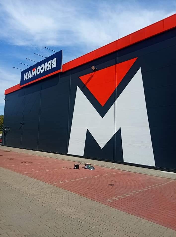 Odświeżona elewacja budynku w Jaworznie z dużym logo w kształcie litery M, widoczne ślady prac malarskich na chodniku.