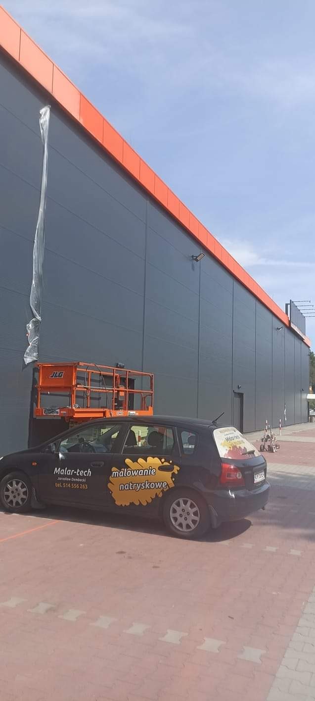 Przygotowanie elewacji budynku przemysłowego do prac malarskich z użyciem podnośnika koszowego, widoczny samochód firmowy z reklamą malowania natryskowego.