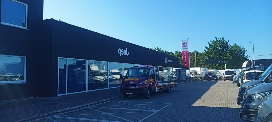 Nowoczesny budynek salonu samochodowego z ciemną elewacją, dużymi oknami wystawowymi i zaparkowanymi pojazdami, w tym lawetą i banerem reklamowym marki.