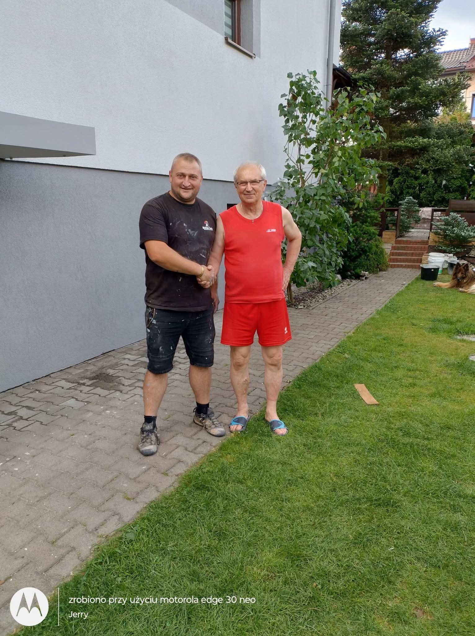 Uścisk dłoni malarza elewacji i klienta na tle świeżo pomalowanej, dwukolorowej ściany domu. Widoczny trawnik i zieleń ogrodu.