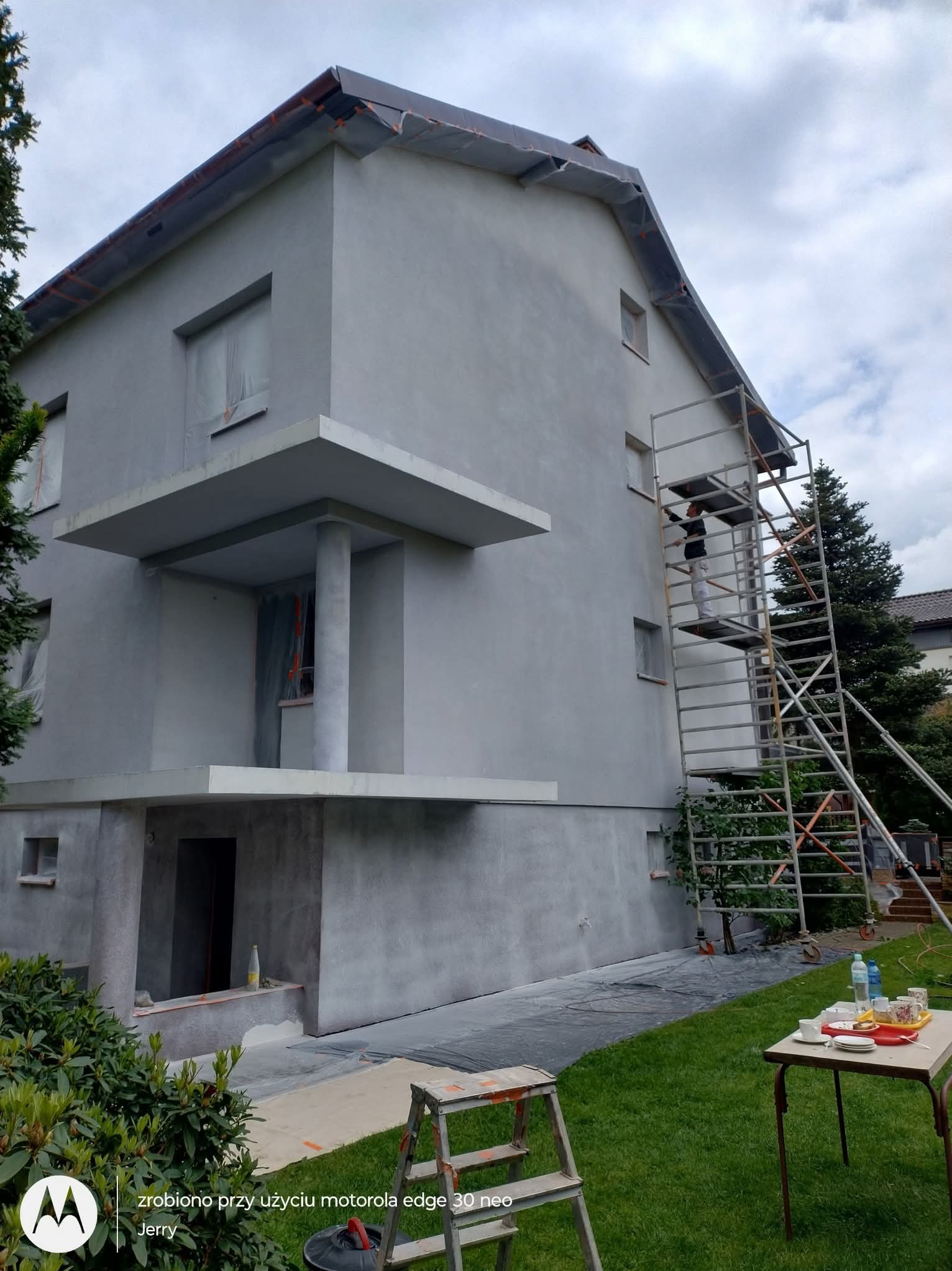 Dwupiętrowy dom w trakcie malowania elewacji na szaro, z rusztowaniem i osobą pracującą na wysokości. Widoczne zabezpieczenia okien i otoczenia.
