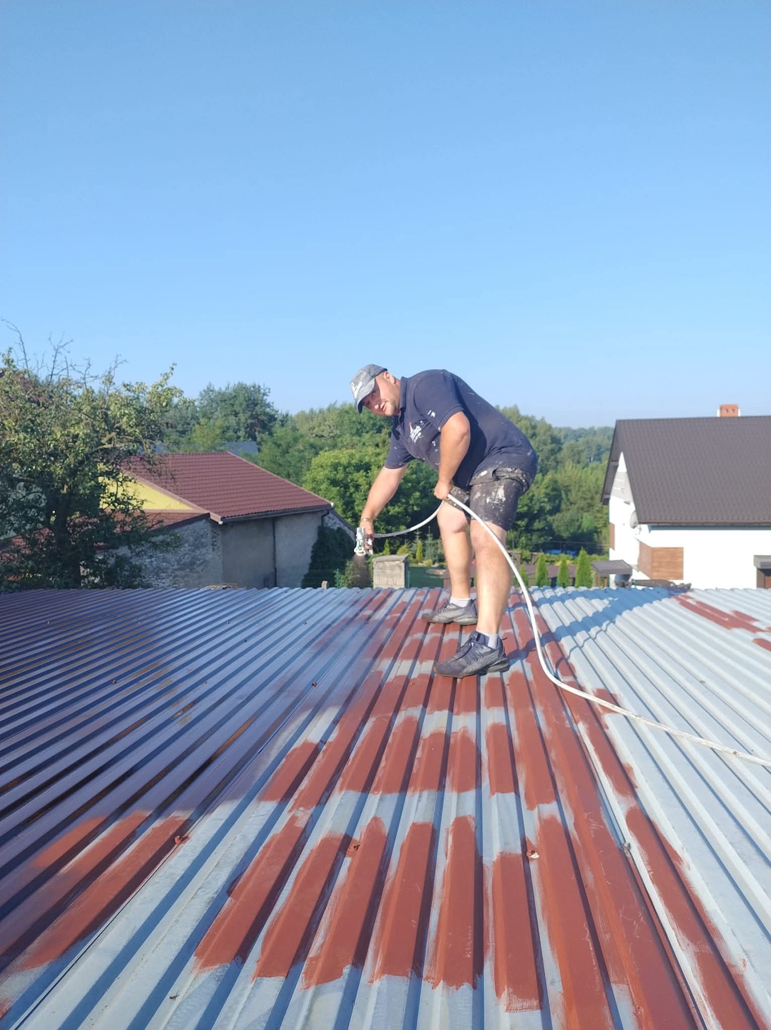 Mężczyzna maluje dach z blachy falistej metodą natryskową na rdzawy kolor. Część dachu już pomalowana, widoczne budynki w tle i błękitne niebo.