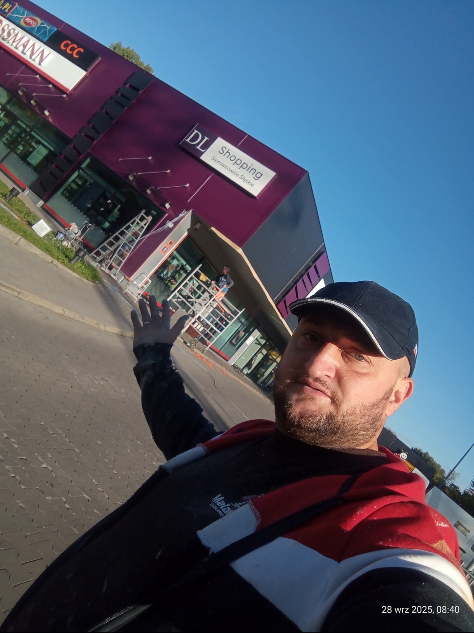 Malowanie elewacji budynku handlowego w fioletowych barwach. Widoczne rusztowania, drabiny i pracownicy podczas pracy. Ujęcie z dołu, z perspektywy osoby stojącej na ziemi.