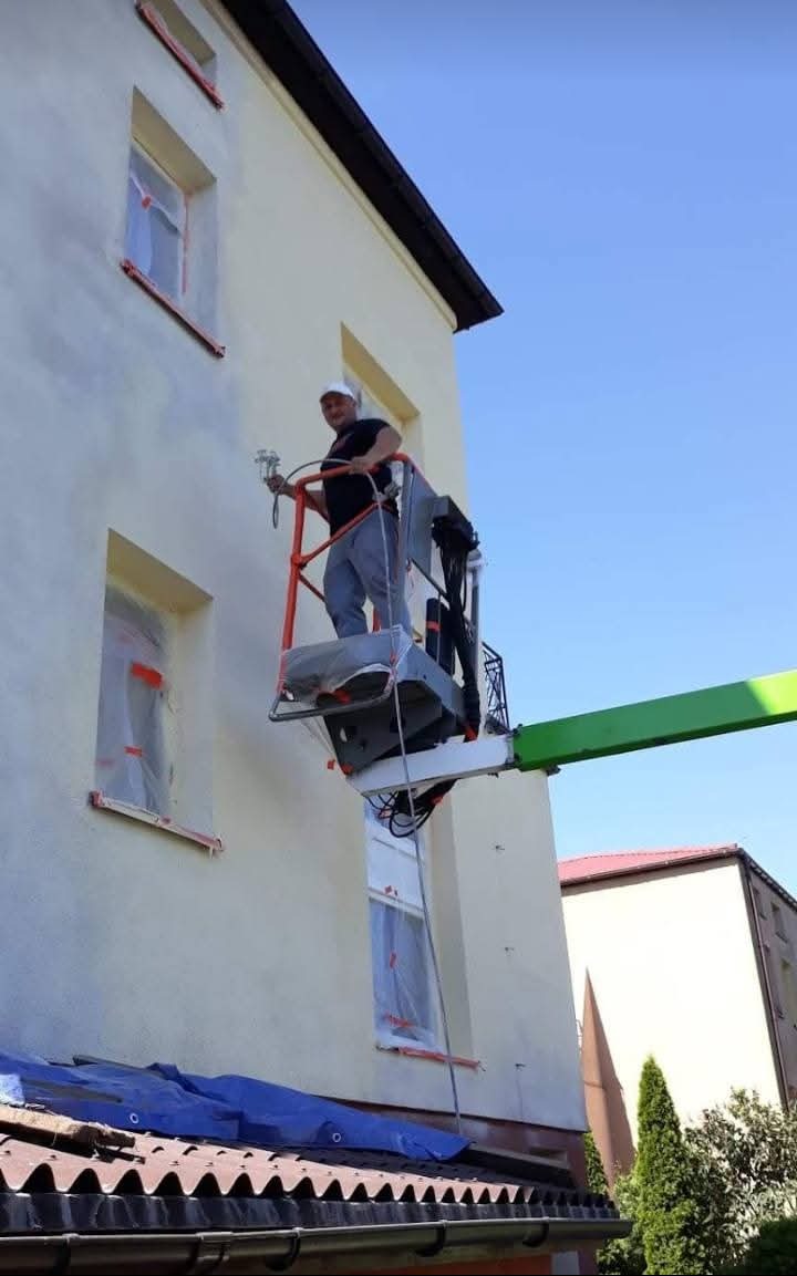 Malowanie elewacji natryskowo z podnośnika koszowego. Okna zabezpieczone folią, widoczny fragment dachu z plandeką. Praca w toku, budynek w jasnych barwach.