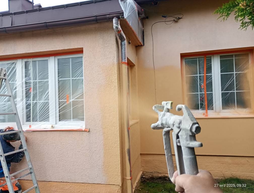 Natryskowe malowanie elewacji domu jednorodzinnego. Okna zabezpieczone folią, widoczny pistolet natryskowy i drabina. Częściowo pomalowana ściana w odcieniach beżu.