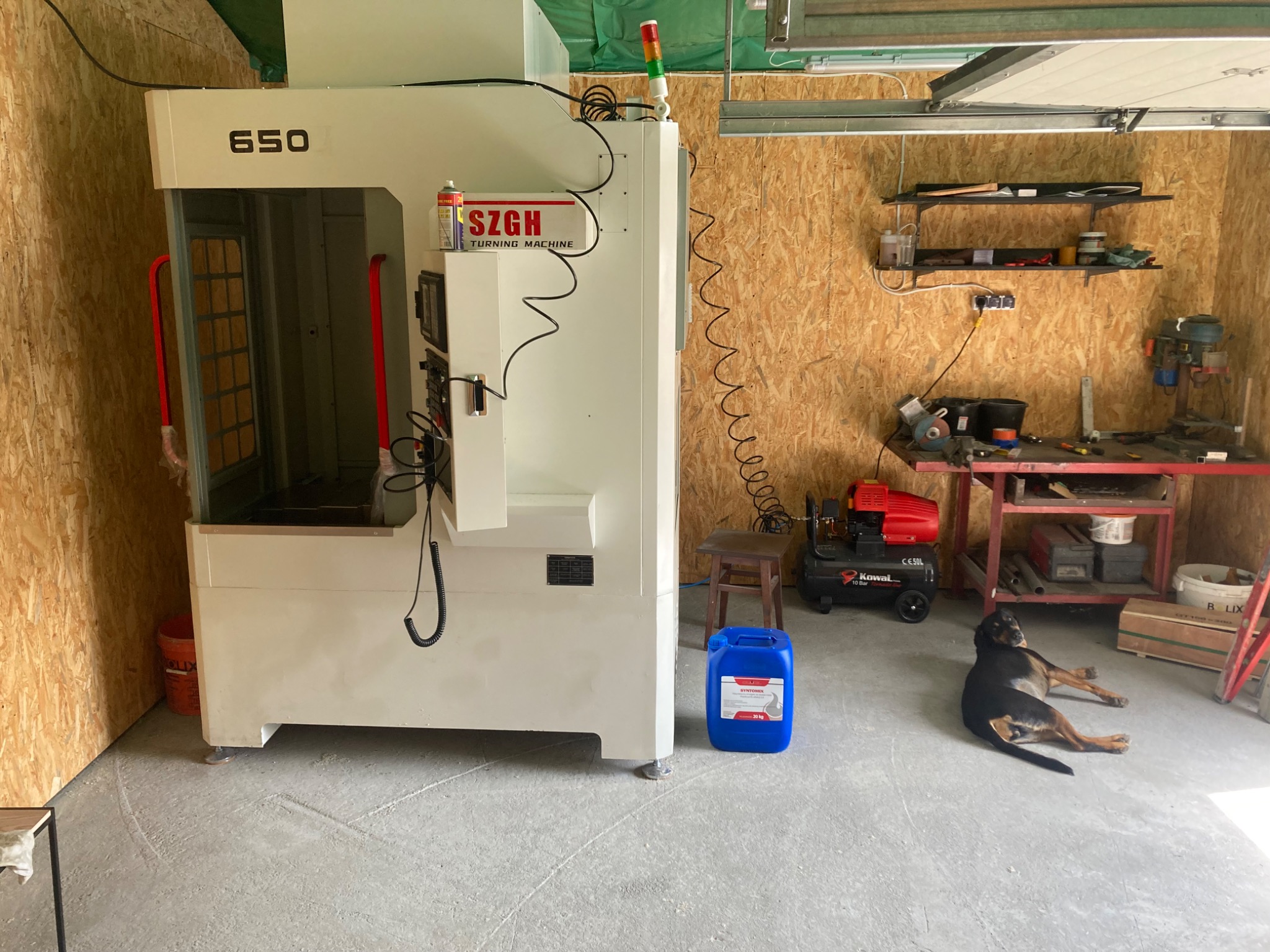 Biała tokarka CNC SZGH 650 w warsztacie z ścianami wyłożonymi płytami OSB, obok leży pies rasy rottweiler.