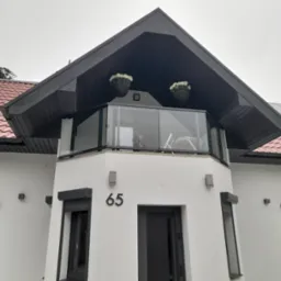 Nowoczesny dom z białymi ścianami i balkonem z przezroczystą balustradą, widoczne kwiaty w donicach na balkonie, rolety zewnętrzne na oknach, numer domu '65' nad wejściem.