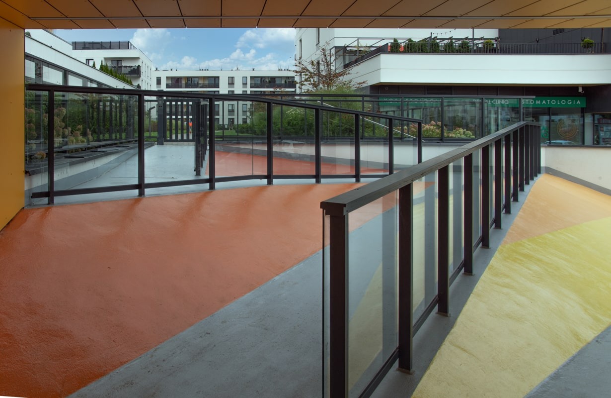 Zewnętrzna przestrzeń z kolorową posadzką i szklaną balustradą z czarnymi profilami, widok na budynki mieszkalne w tle.