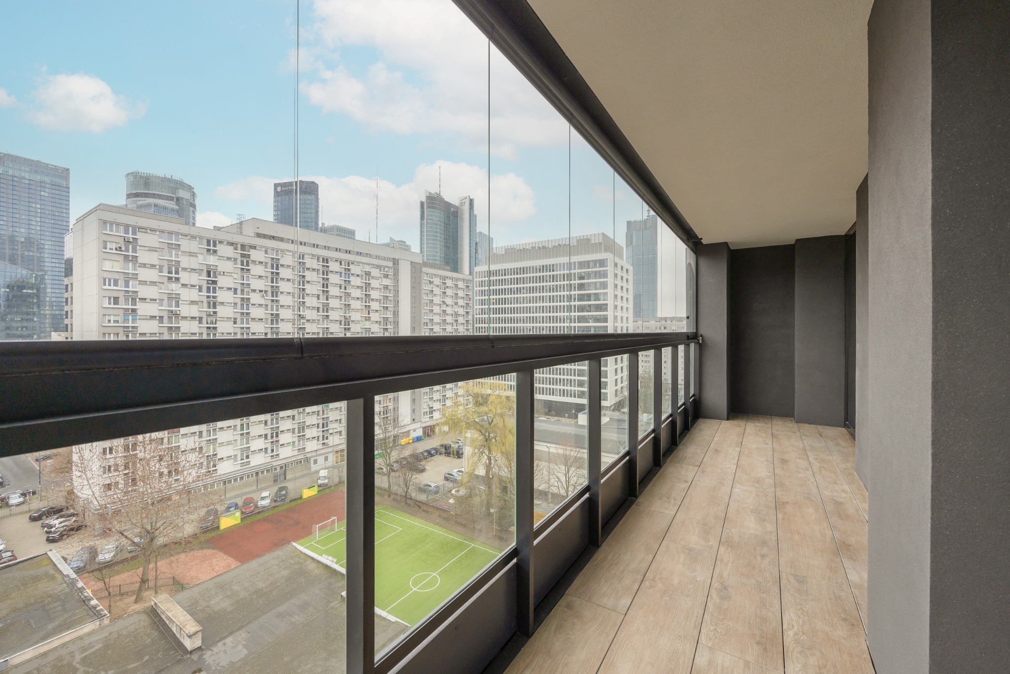 Szklana balustrada balkonowa z widokiem na panoramę miasta i boisko sportowe, drewnopodobna podłoga, minimalistyczny design.