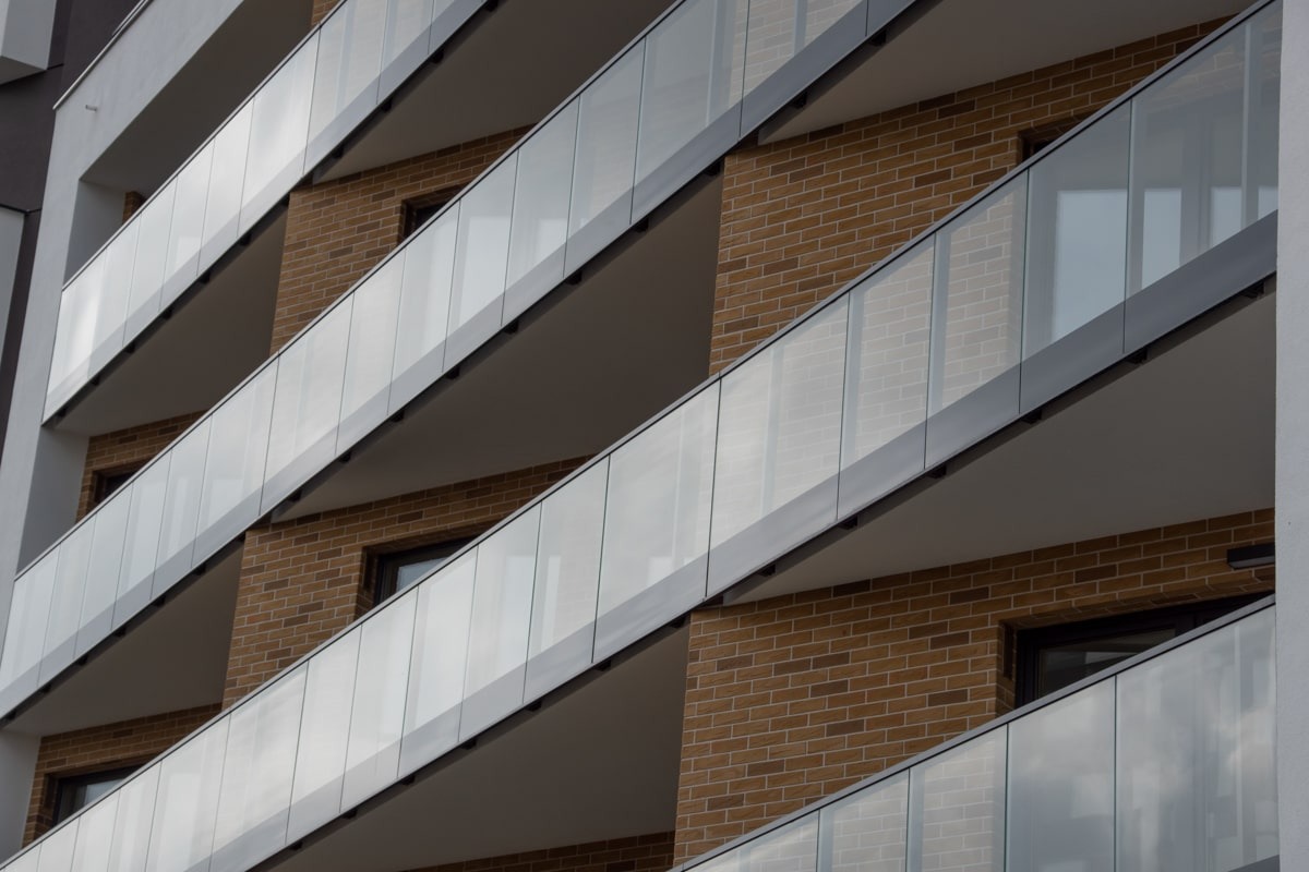 Elewacja budynku z balkonami zabezpieczonymi przezroczystymi taflami szkła, widoczne ceglane wykończenie ścian i szara konstrukcja wsporcza.