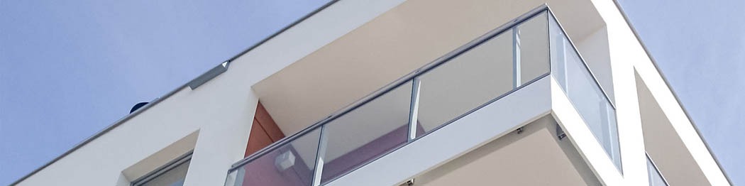 Nowoczesny balkon z minimalistyczną szklaną balustradą, widok z dołu na tle błękitnego nieba, biała elewacja budynku z elementami drewnianymi.