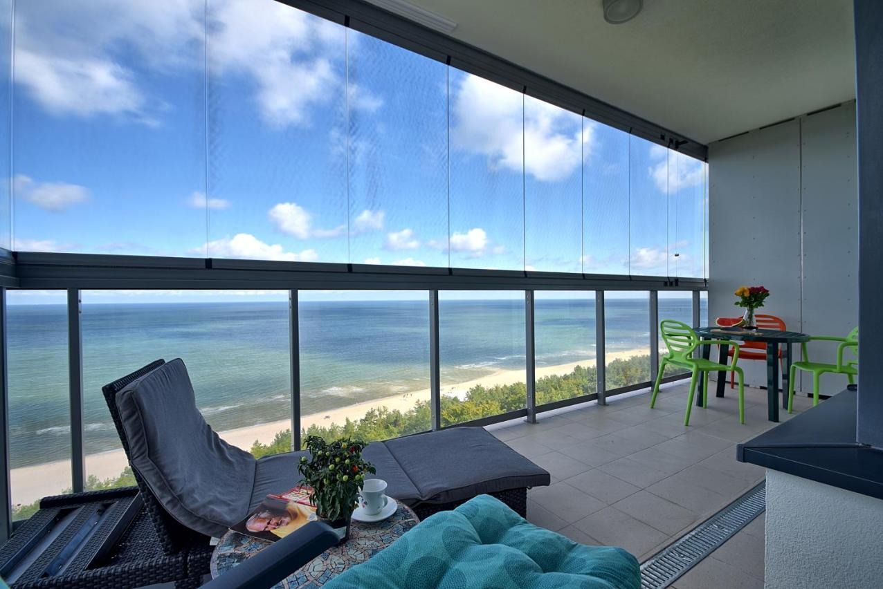 Zamykany balkon z widokiem na morze, szklane panele bezramowe, meble wypoczynkowe i stół z krzesłami.