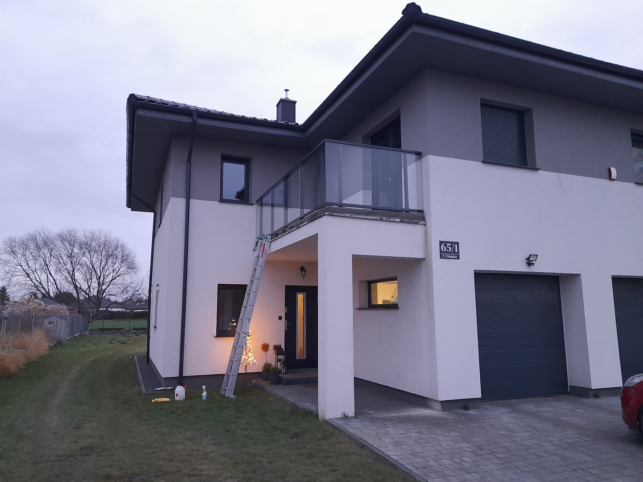 Nowoczesny dom dwukondygnacyjny z balkonem zabezpieczonym szklaną balustradą, z drabiną opartą o ścianę i dekoracyjnym drzewkiem przed wejściem, sfotografowany o zmierzchu.