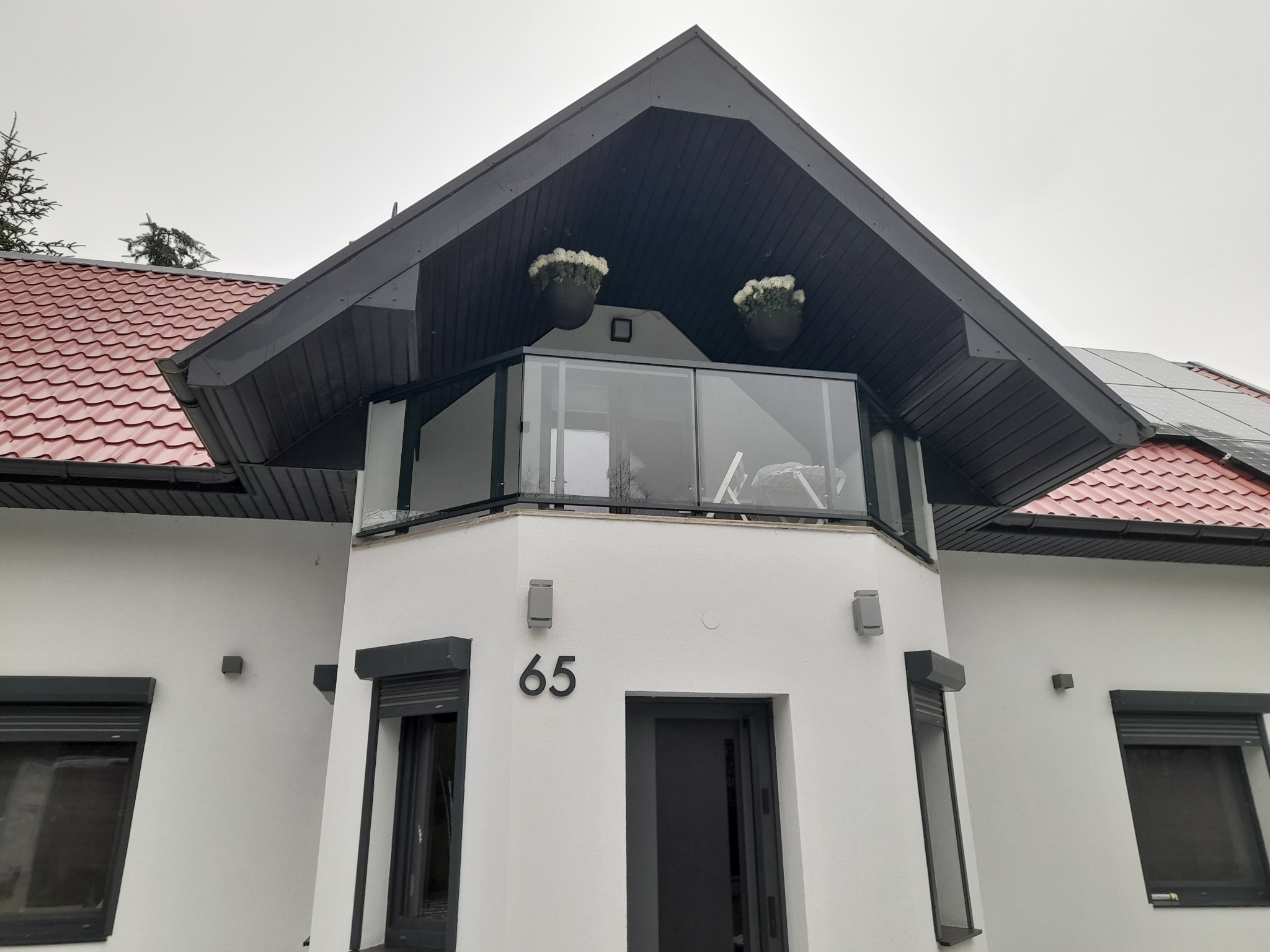 Nowoczesny dom z białymi ścianami i balkonem z przezroczystą balustradą, widoczne kwiaty w donicach na balkonie, rolety zewnętrzne na oknach, numer domu '65' nad wejściem.