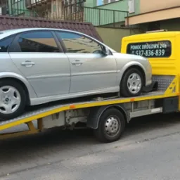 Srebrny Opel Vectra na żółtej lawecie na tle miejskiej ulicy, widoczny numer telefonu 'Pomoc Drogowa 24h'