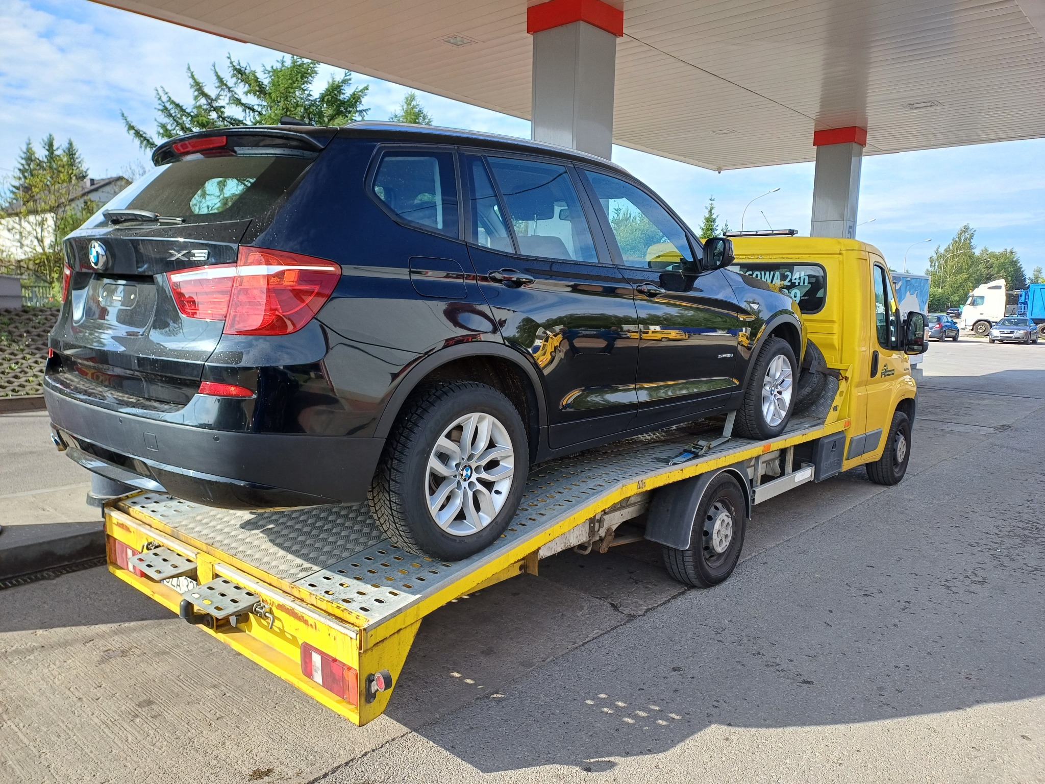 Żółta laweta z czarnym BMW X3 na platformie, zaparkowana pod zadaszeniem stacji benzynowej.