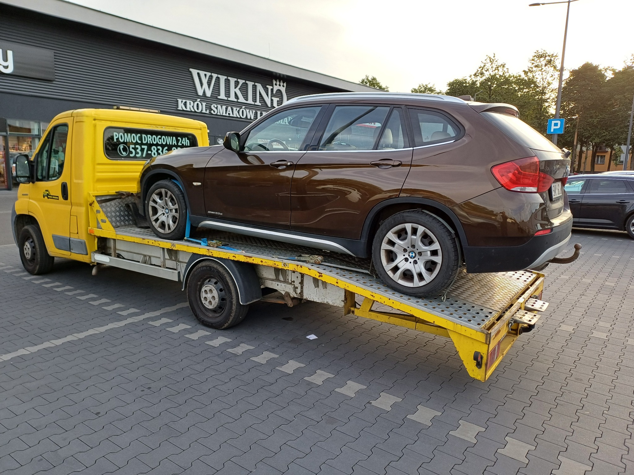Brązowe BMW X1 na żółtej lawecie pomocy drogowej z numerem telefonu, zaparkowanej na parkingu przed sklepem Wiking Król Smaków.